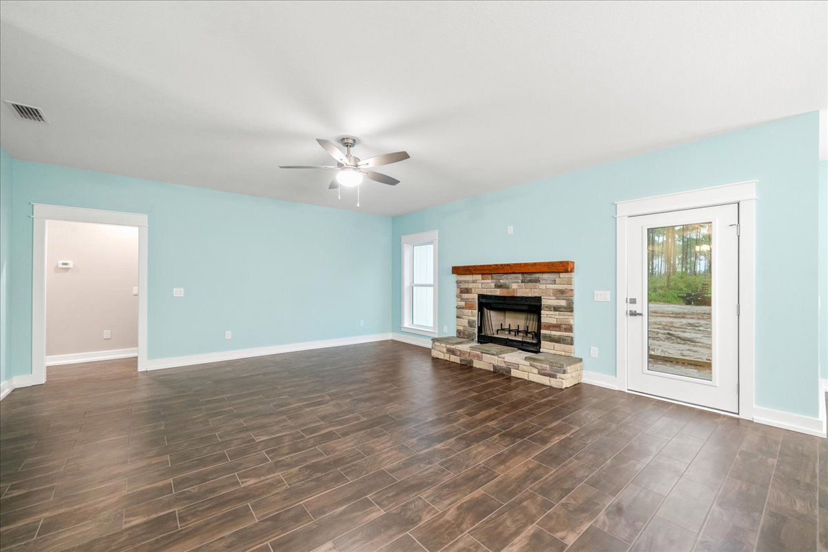 Living room with laminate wood flooring, white walls, black-framed fireplace with wood mantel, ceiling fan with light, white door opening to forest view, visible thermostat on wall