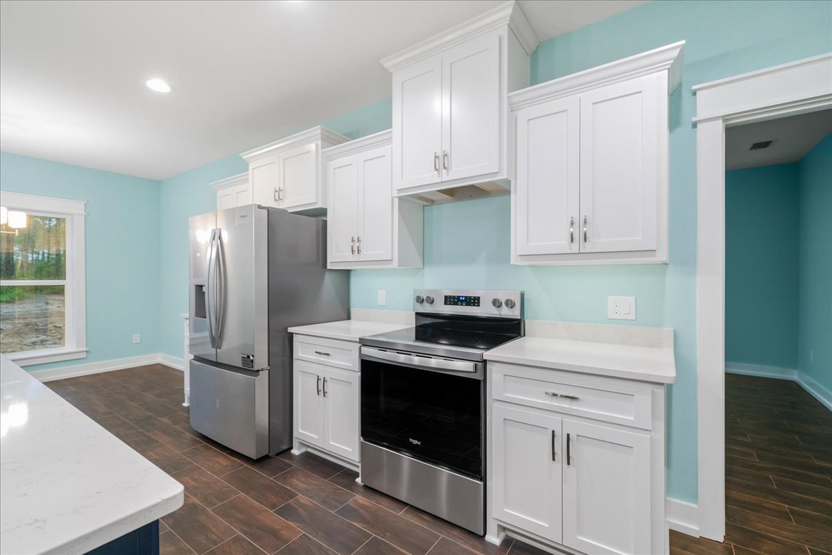 White kitchen cabinets with silver handles, stainless steel refrigerator, black stove, light countertop, white-framed window, and sink.