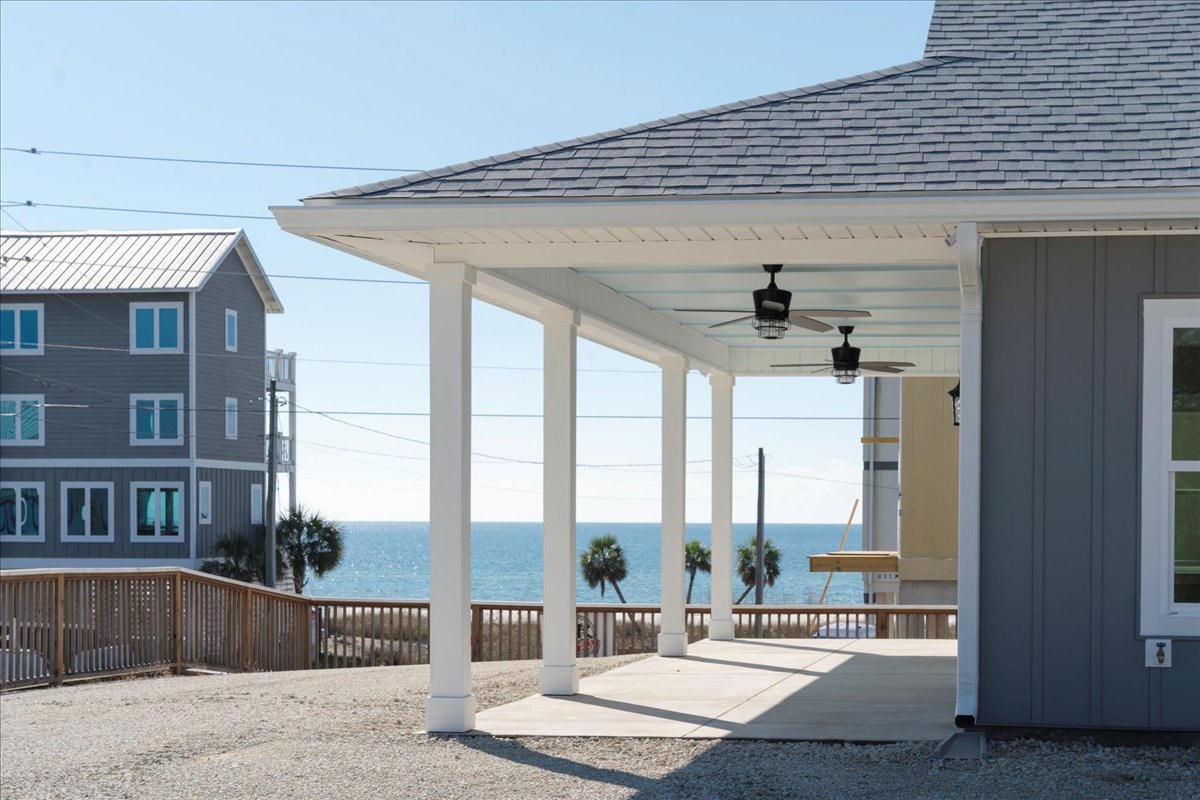 Grey house with white trim featuring a covered porch with ceiling fan, concrete walkway bordered by white pillars, window overlooking green foliage, wooden fence with post, and