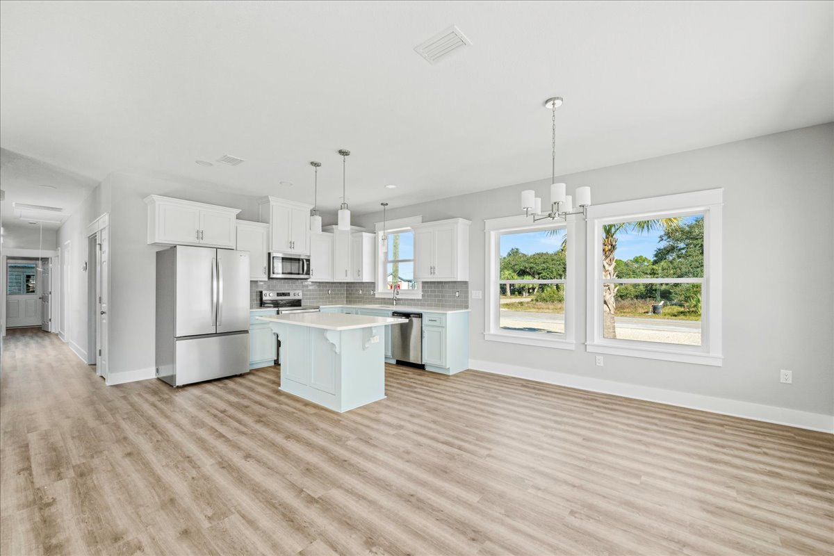 Kitchen with light wood flooring, white cabinetry, white island, stainless steel refrigerator, and large window overlooking trees and a road