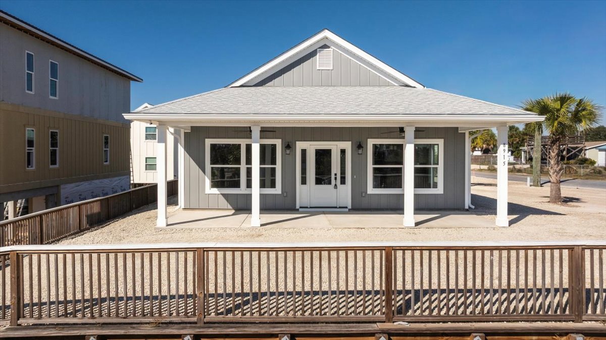 Grey siding house with white pillars, wooden porch railing casting shadow, white framed window, glass panel door, palm tree in landscaped front yard.