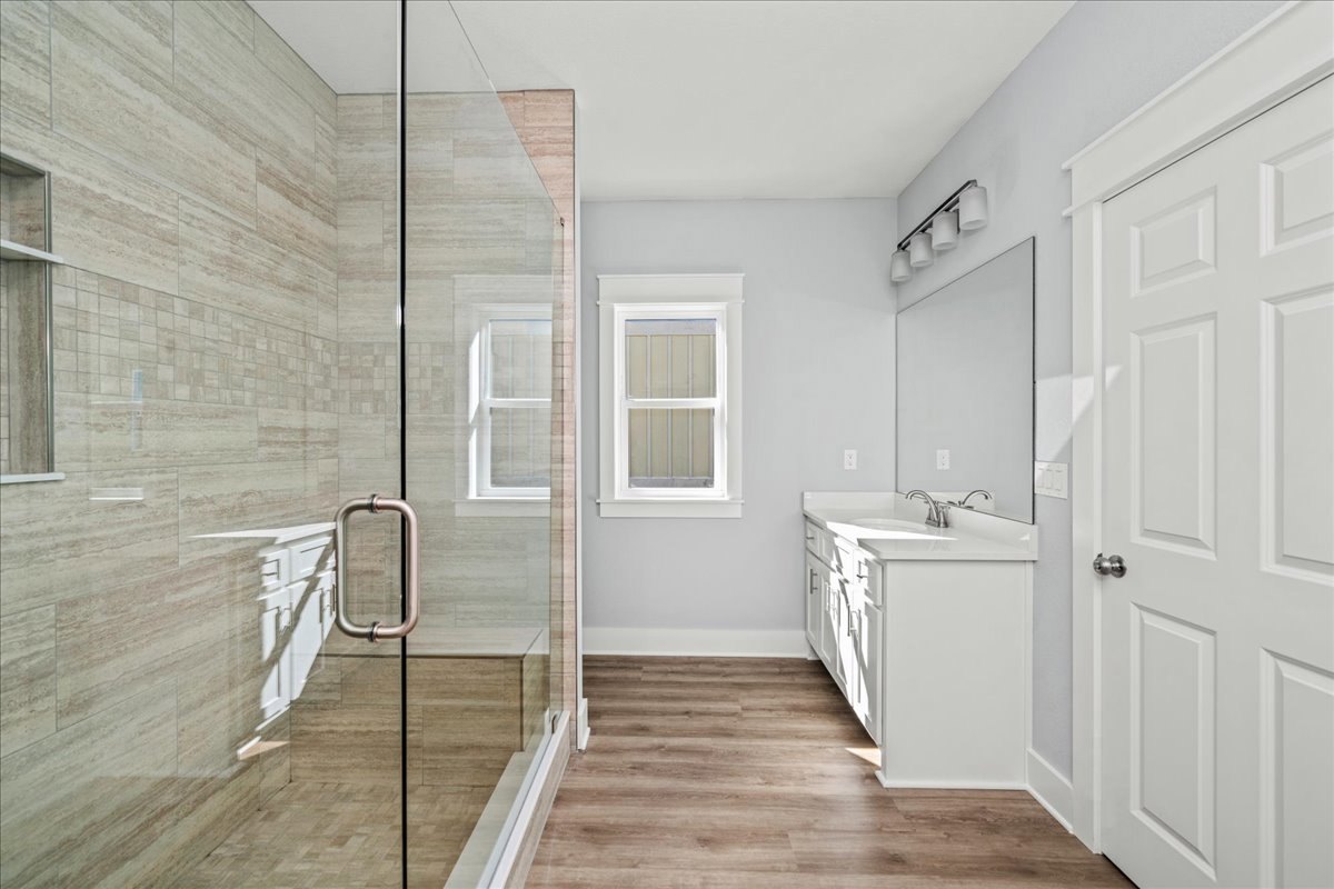 Bathroom with glass shower enclosure, white framed window, wood flooring, white walls, white sink and mirror, four-shade light fixture, and partial view of white door