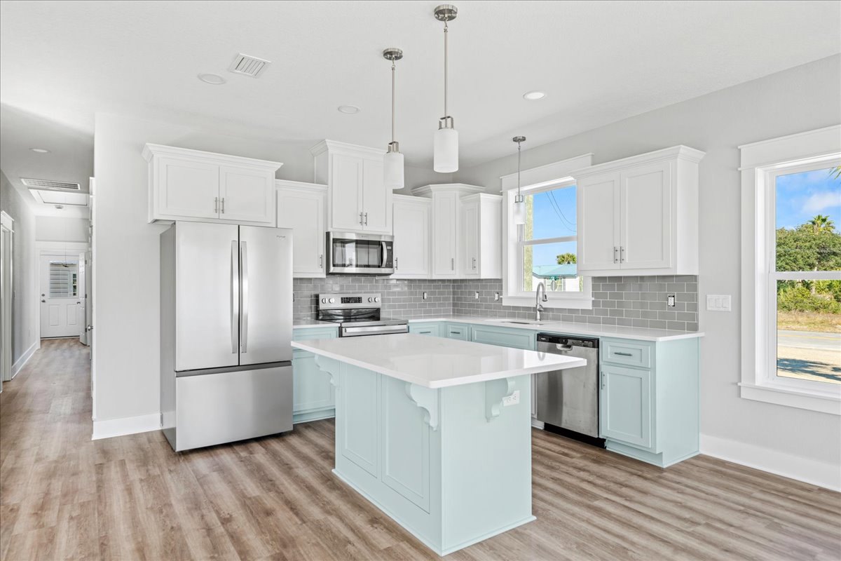 White kitchen with shaker cabinets, central white island, wood flooring, stainless refrigerator, silver sink set in white countertop, large window framing trees and blue sky
