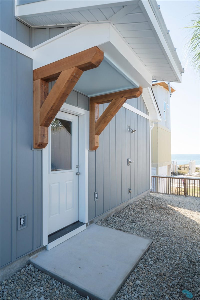 Modern home exterior with white walls, black-framed windows, wooden front door, covered porch supported by wooden beams, concrete slab walkway, gravel landscaping, wooden fence and
