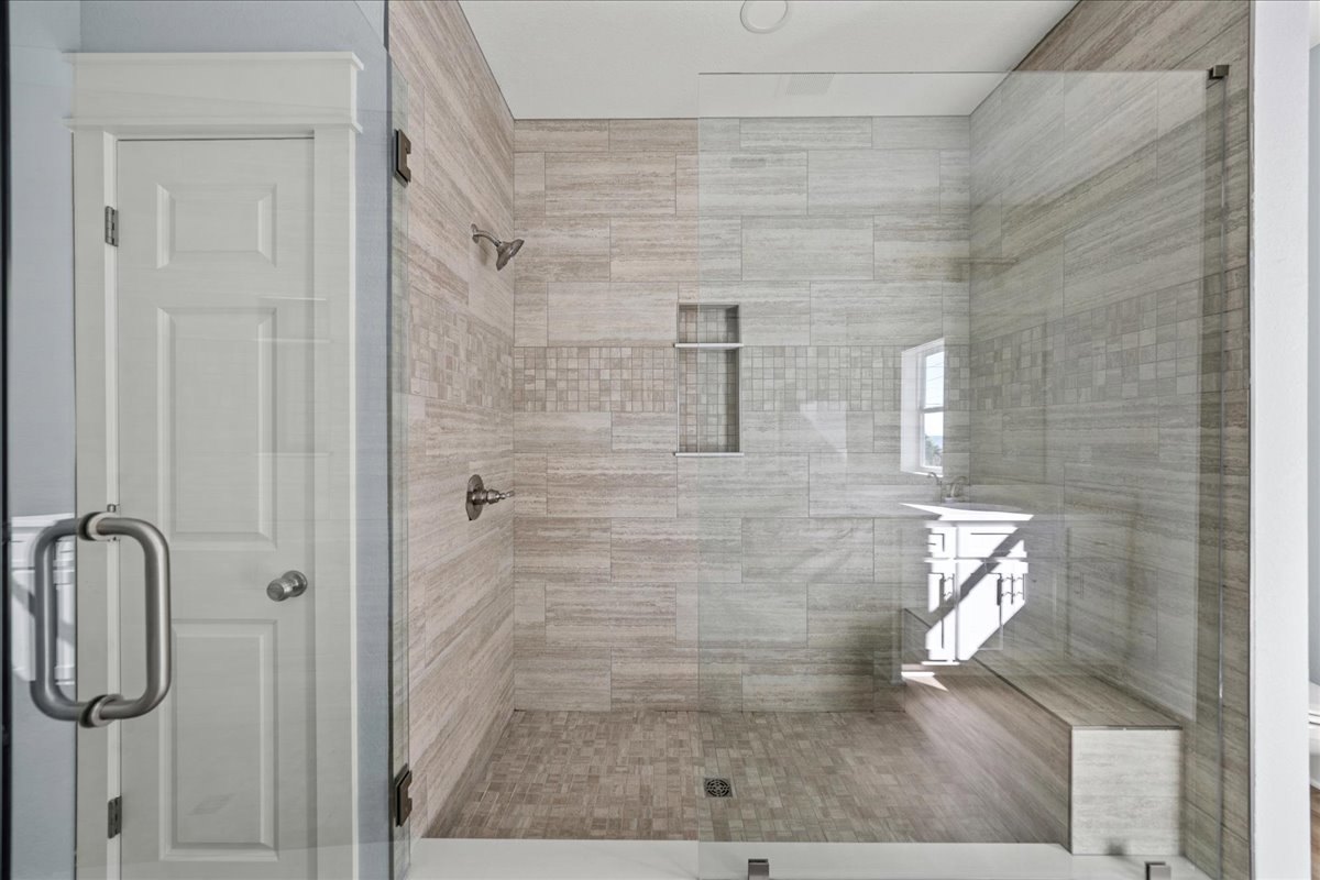 Glass shower enclosure with tile floor, built-in bench, and adjacent white door featuring a silver handle; window with white frame allows natural light into the bathroom.