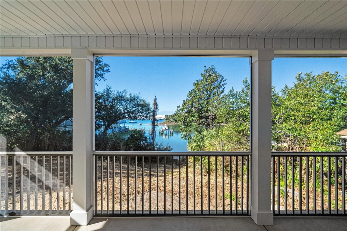 Covered porch with black metal railing overlooking a lake, trees, and open field under a blue sky