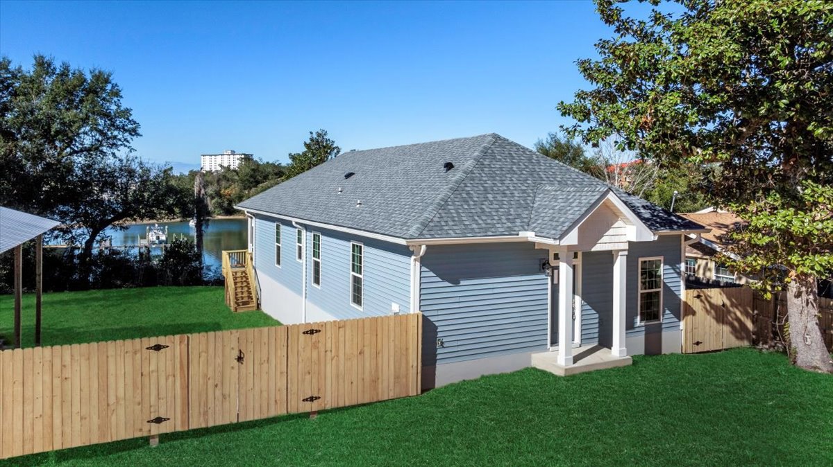 Two-story home with gray siding, wooden privacy fence, grassy yard, and mature tree in the background; exterior wooden stairs lead to a side entrance.