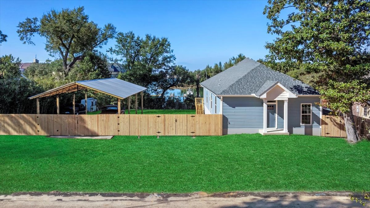 Two-story house with blue roof, surrounded by green grass lawn, wooden fence, and mature tree in the background