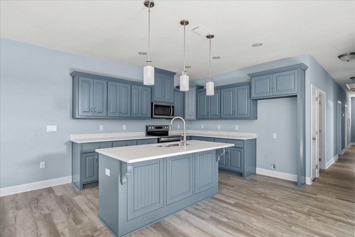 Spacious kitchen featuring a large central island with built-in sink and white faucet, stainless steel stove with matching hood, white cabinetry, stone countertops, and hardwood