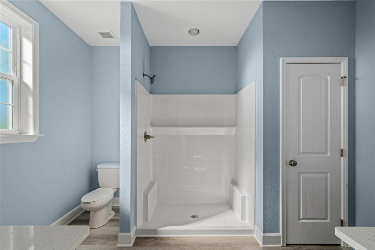 Bathroom with white tile shower, silver faucet, white toilet, white door with silver doorknob, and window letting in natural light