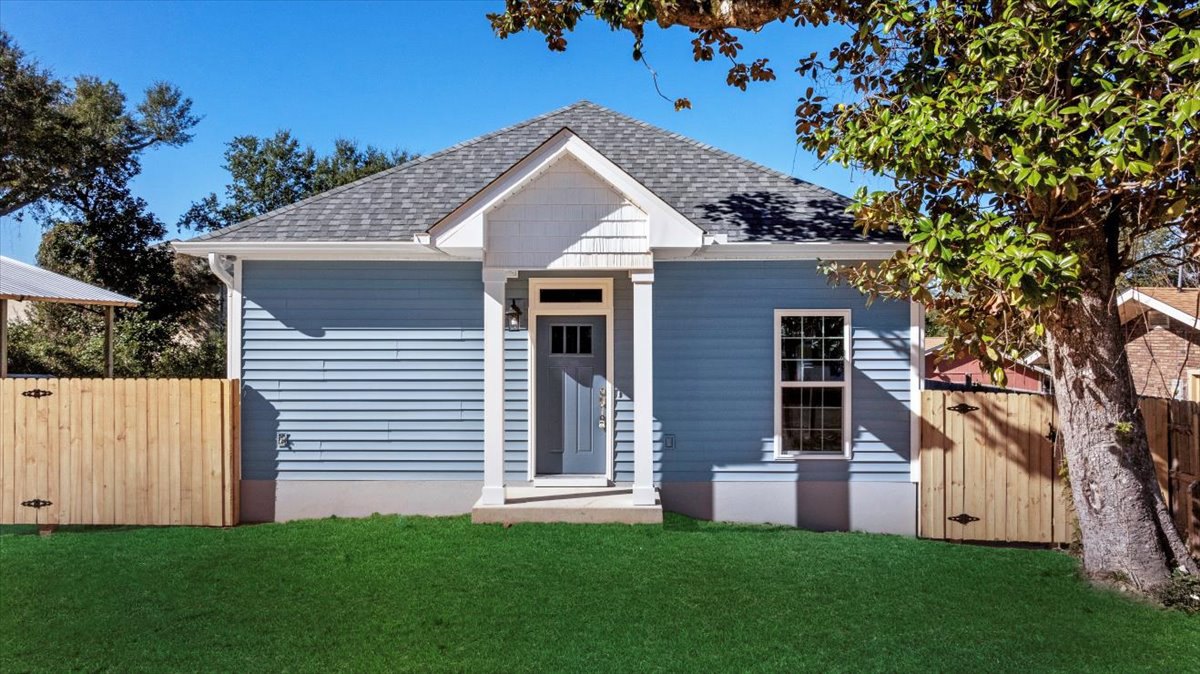 Blue siding home with white trim and door, multi-pane windows, wooden fence with metal hinges, green lawn, mature trees, and person standing on grass.