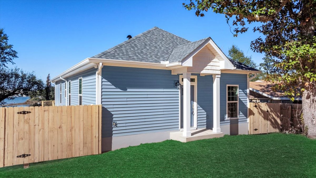 Blue siding house with multi-pane windows, white front door, wooden fence with gate, green grass lawn, leafy tree, and clear blue sky