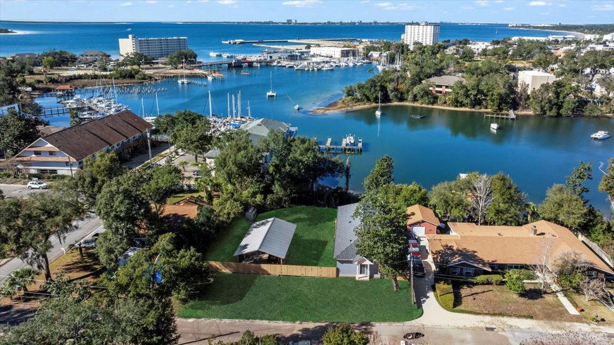 Lakeside custom home with white exterior, multiple windows, sloped roof, surrounded by trees, boats docked along shoreline, neighboring buildings visible across water under blue