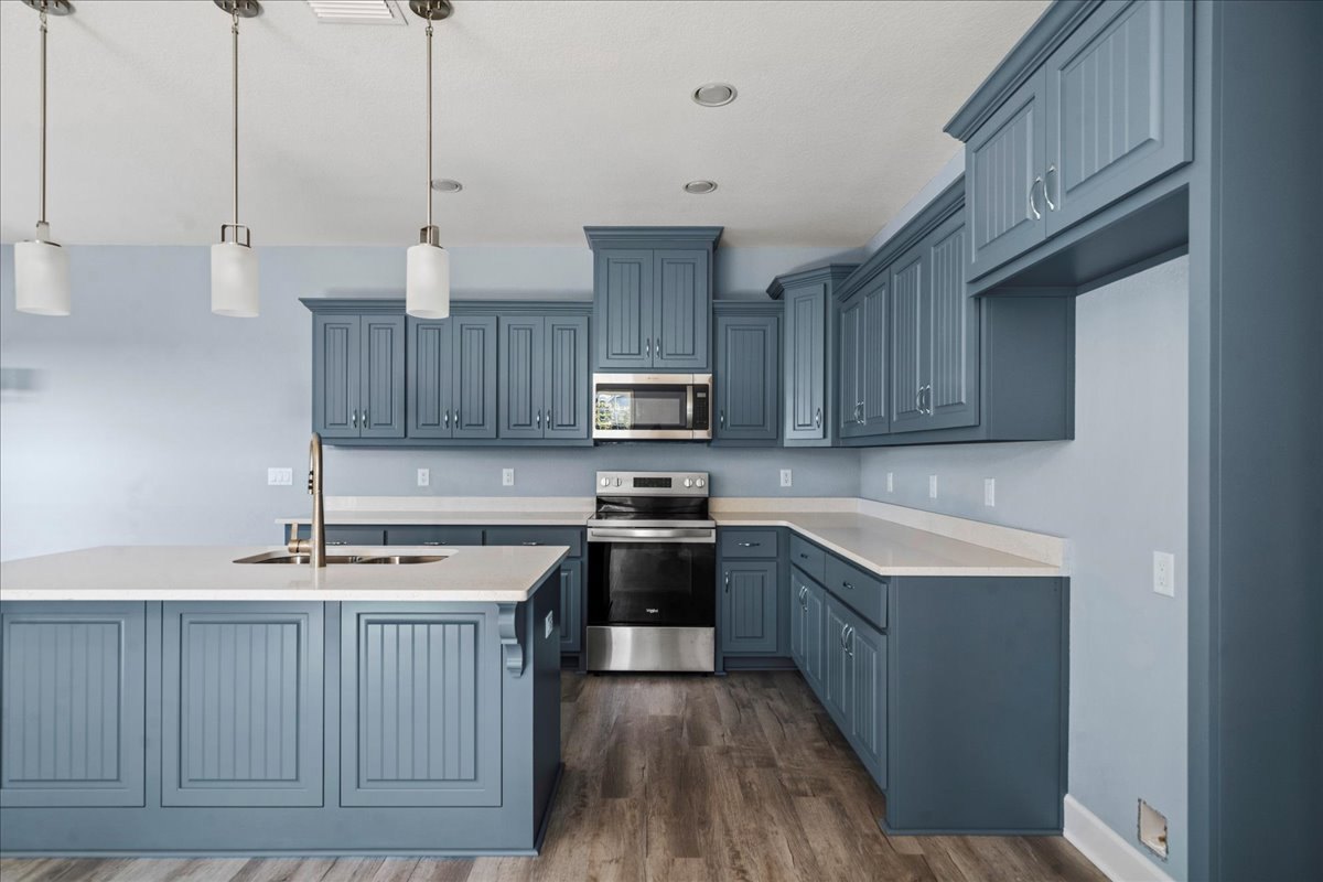Blue shaker cabinets with brushed nickel hardware, white quartz countertops, stainless steel oven and microwave, undermount sink, glass bottle on counter, white cylindrical pendant