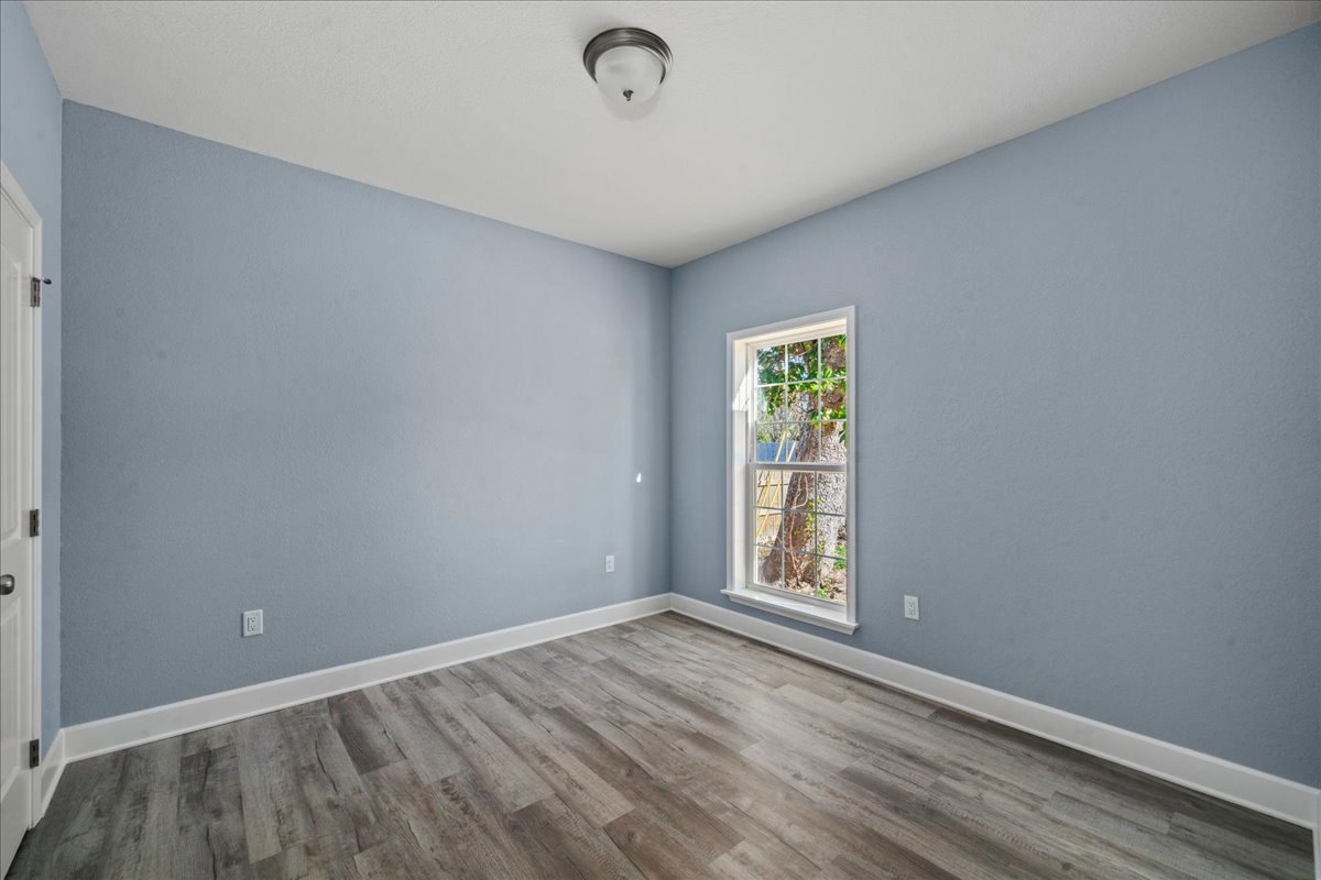 Light blue walls, wide window with view of tree, wood flooring, ceiling light fixture, white door with visible hinge