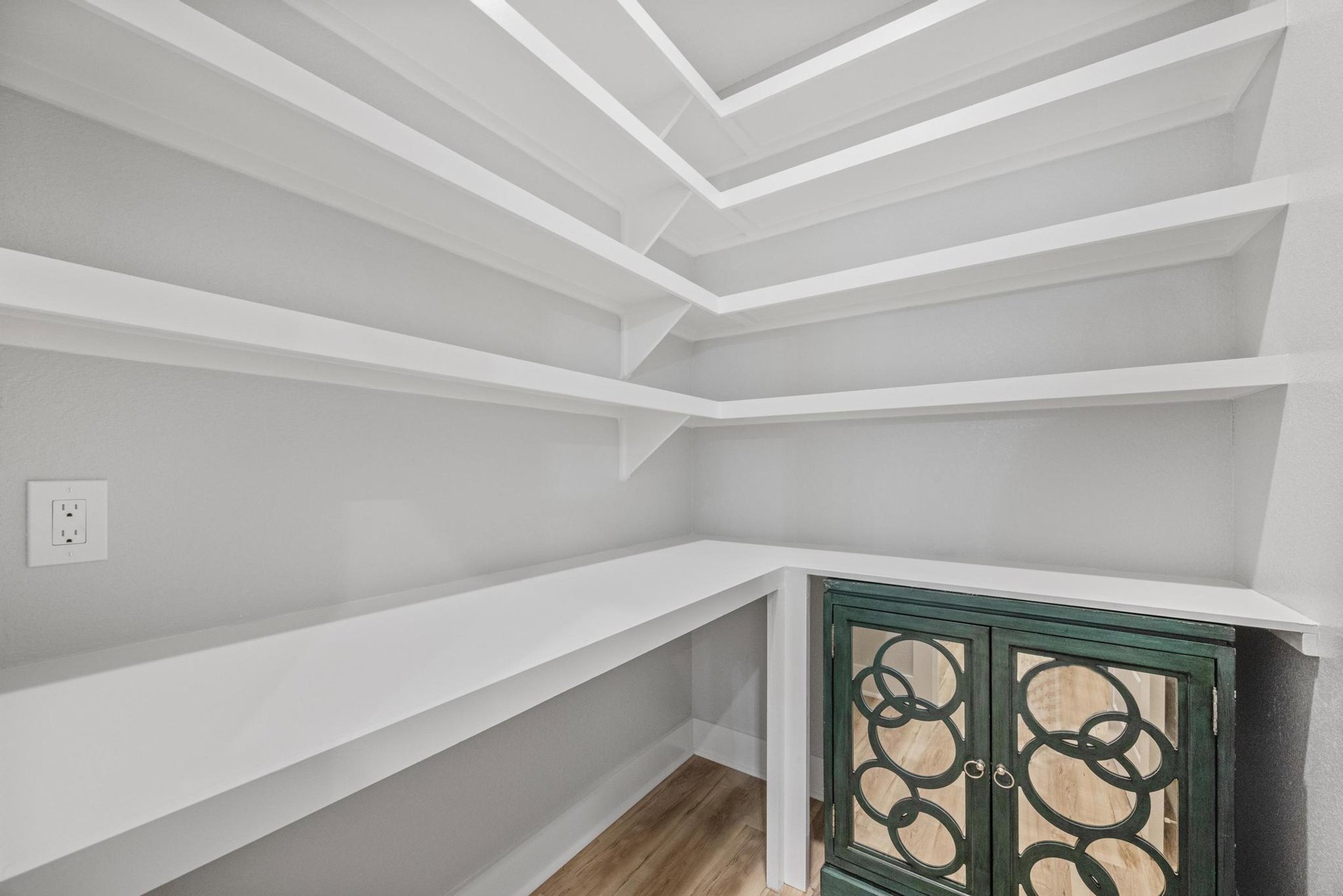 Built-in wood shelves and a green cabinet with marble countertop, white ceiling beams, hardwood flooring, and a white electrical outlet on the wall