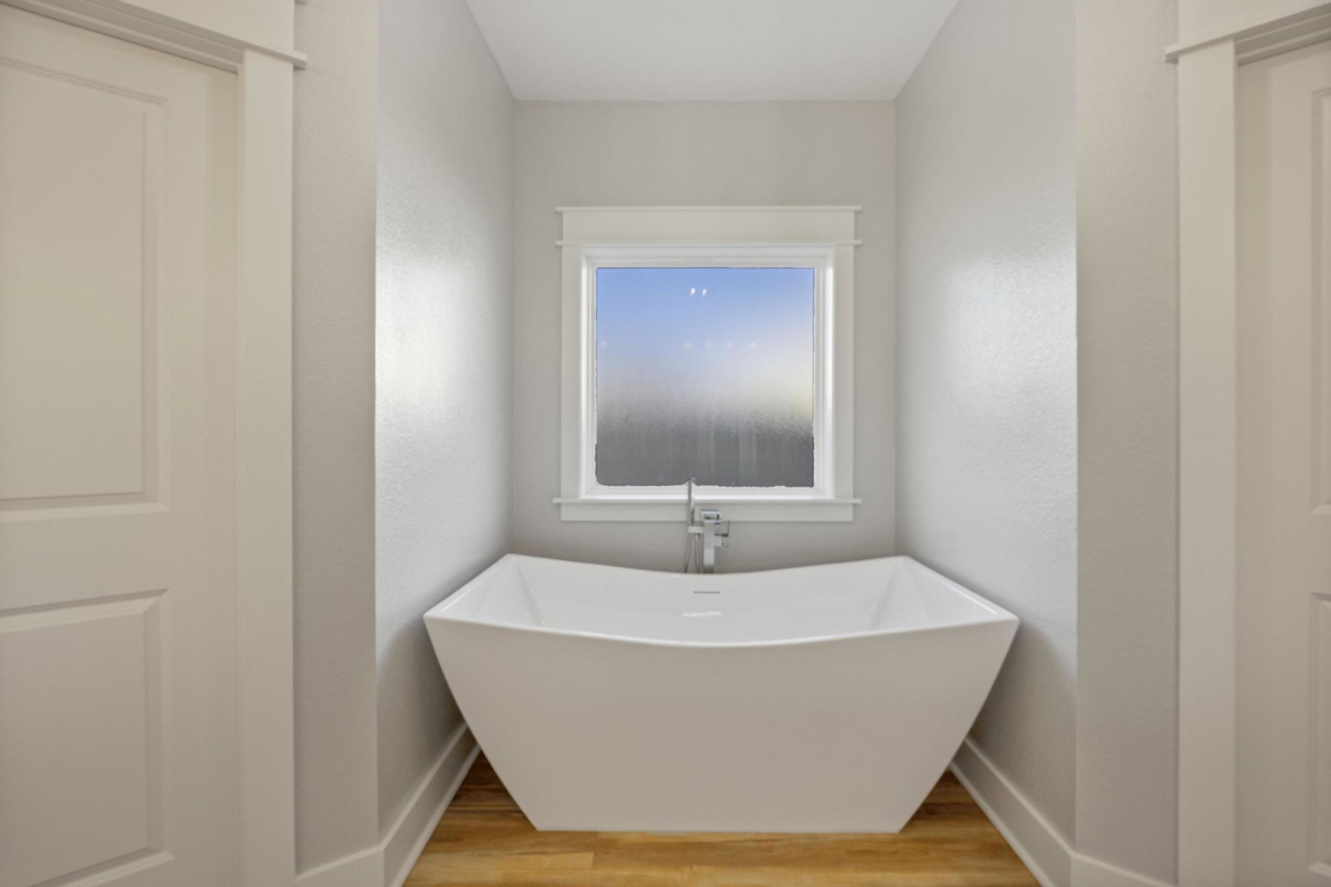 Freestanding white bathtub with metal handle beside frosted glass window, white door, wood surface, and tiled walls; blurred shower head visible in background.