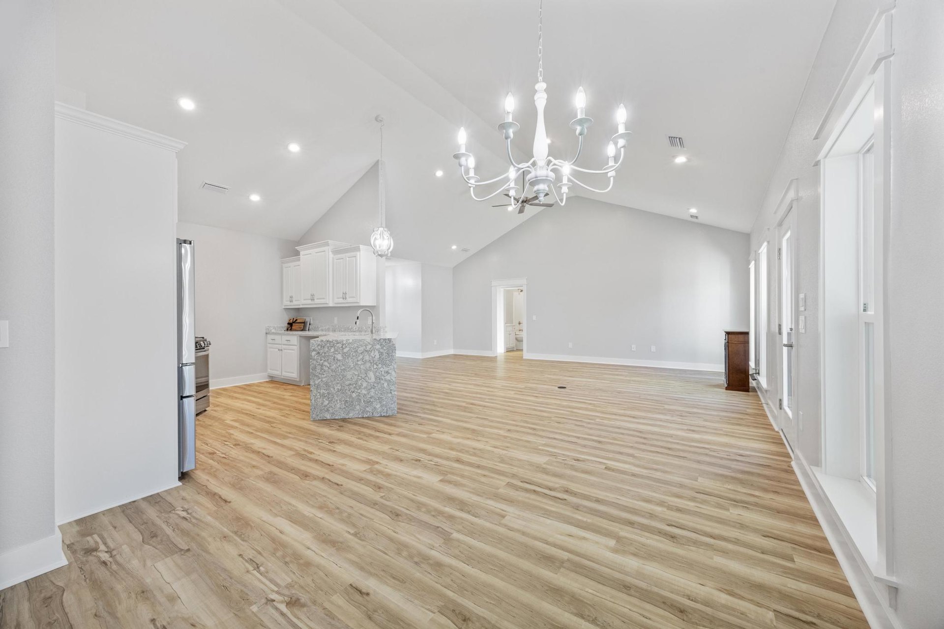 Open-concept kitchen with marble countertops, wood flooring, white cabinetry with silver handles, and a white chandelier hanging from the ceiling