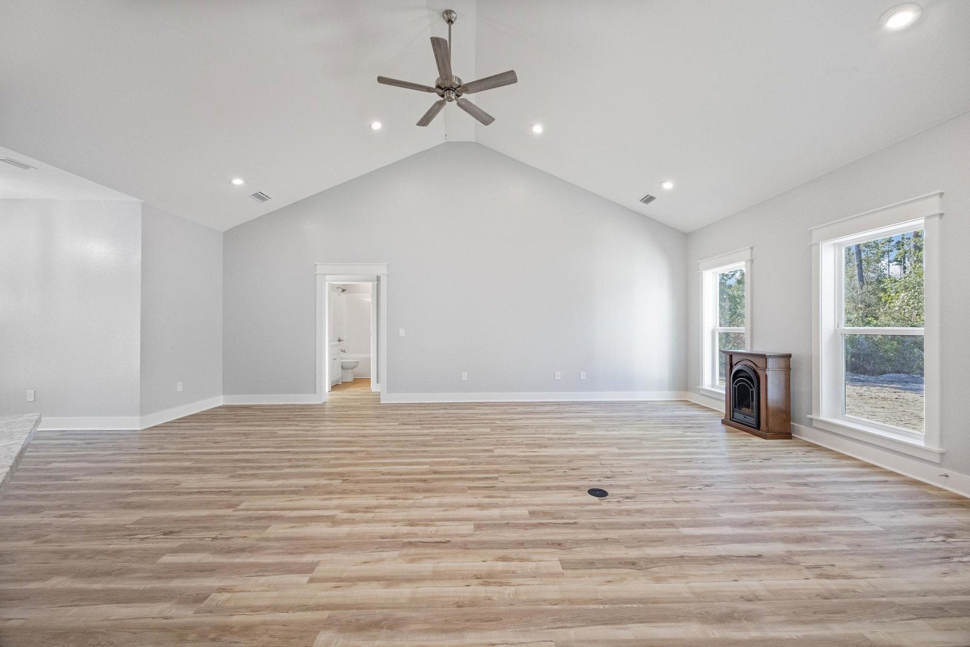 Spacious living area with wood flooring, white walls, ceiling fan with light fixture, fireplace, large window overlooking trees, and adjacent white bathroom with toilet and sink