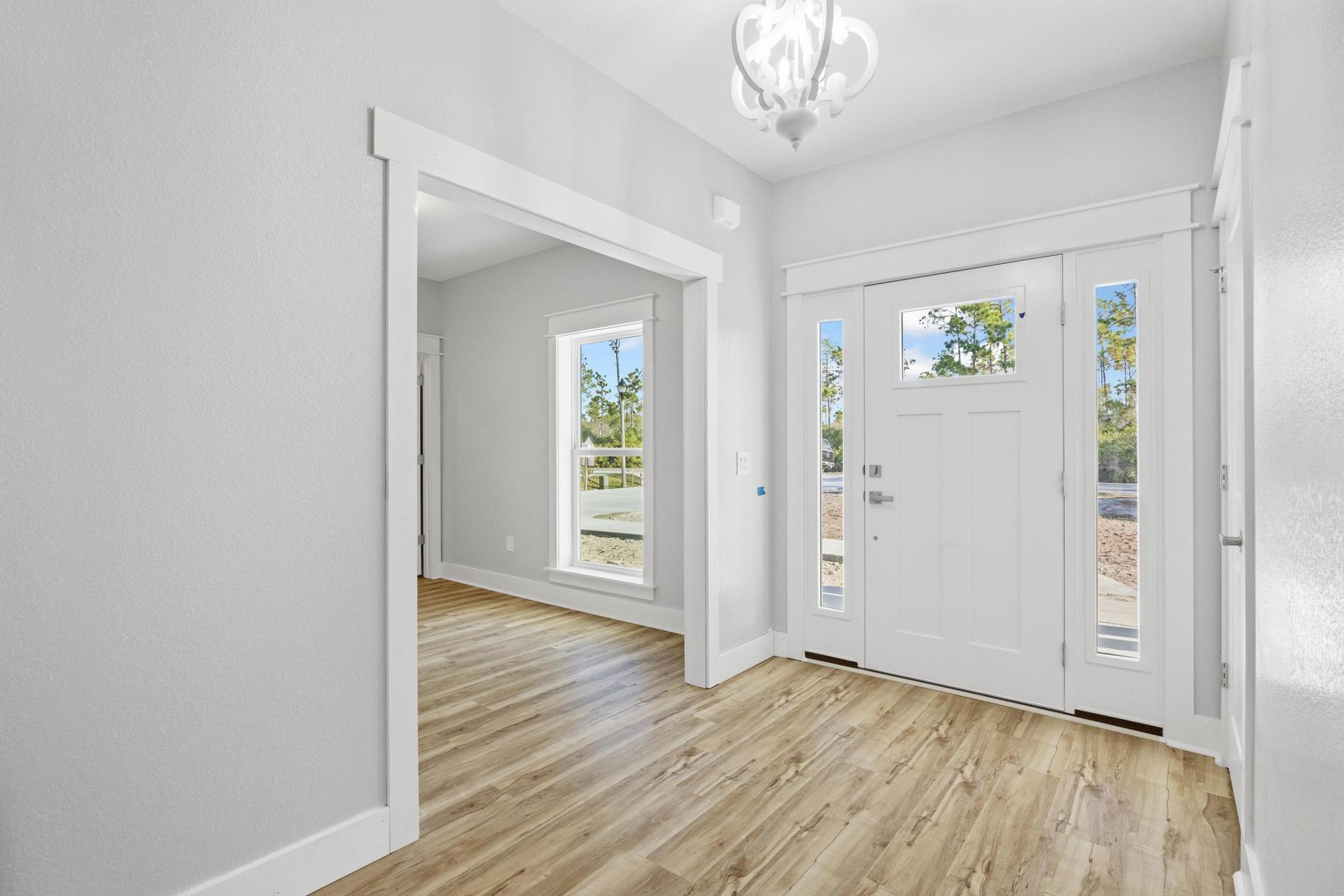 White room with wood flooring, white plaster walls, glass-paneled white door, chandelier overhead, large window revealing trees and streetlight outside.