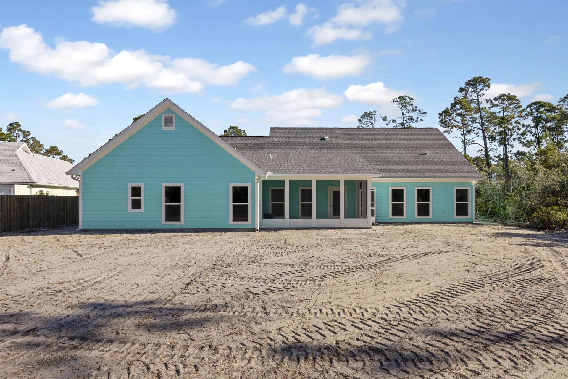 Blue house with white pillars and window frames, large sandy yard with visible tire tracks, wooden fence along the side, tree in the background under a partly cloudy sky.