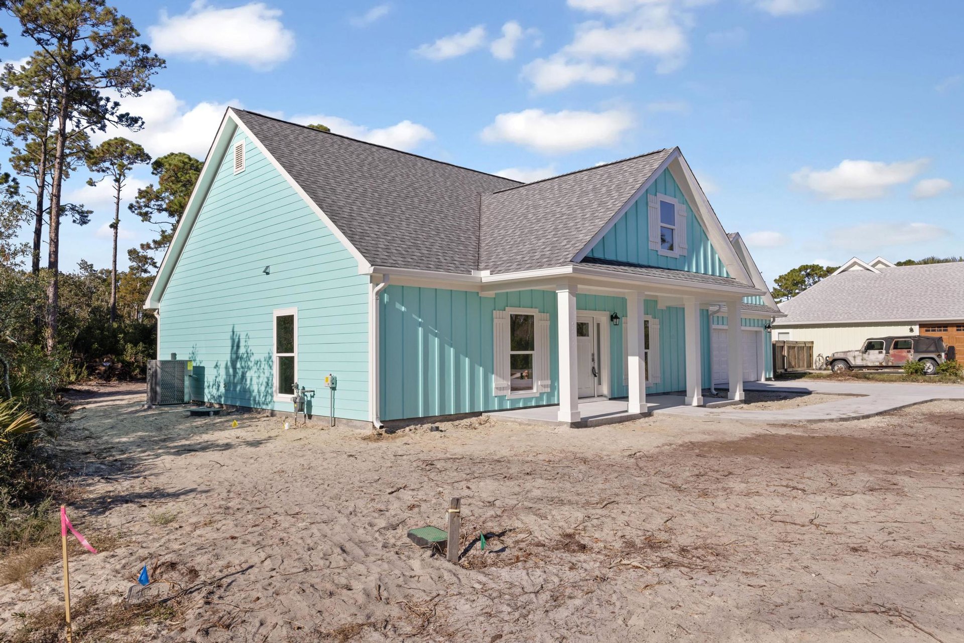 Blue house with white columns and window frames, dirt yard with a hole in the ground, rusted car parked nearby, tall trees in the background, cloudy sky overhead.