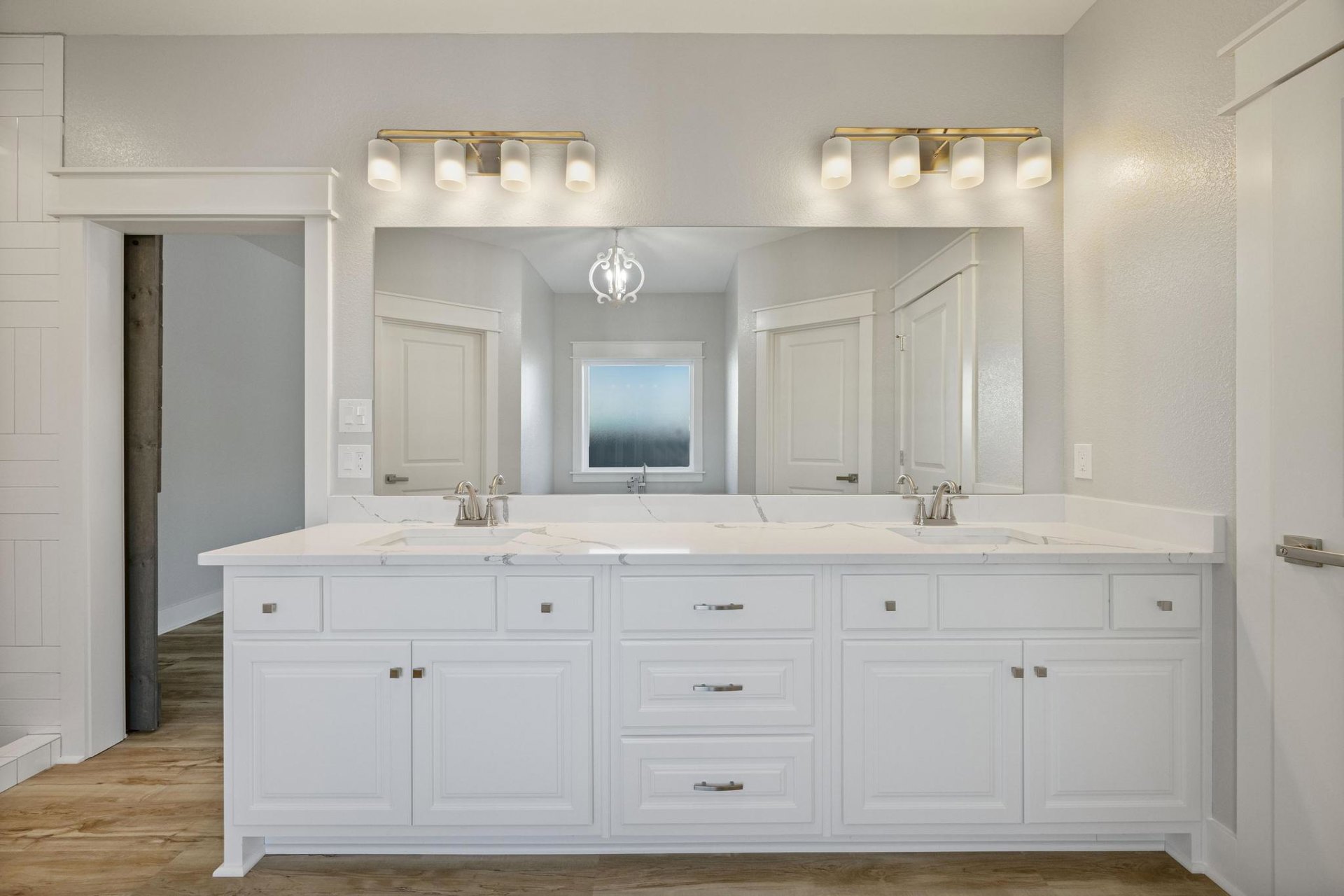 Bathroom with expansive mirror above white cabinetry, frosted glass window, white countertop with silver handles, and dual-lamp light fixture.