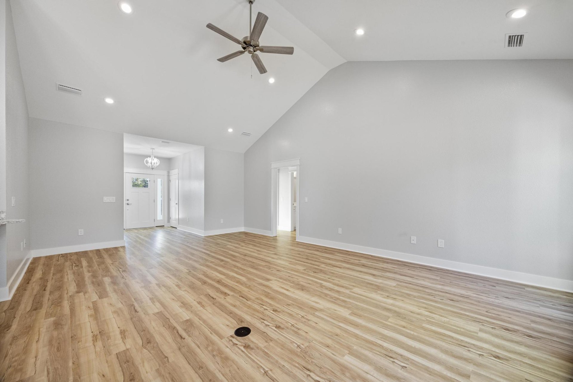 Spacious room with wood flooring, ceiling fan with wood blades, white chandelier, white door with window and light switch, plaster walls and ceiling