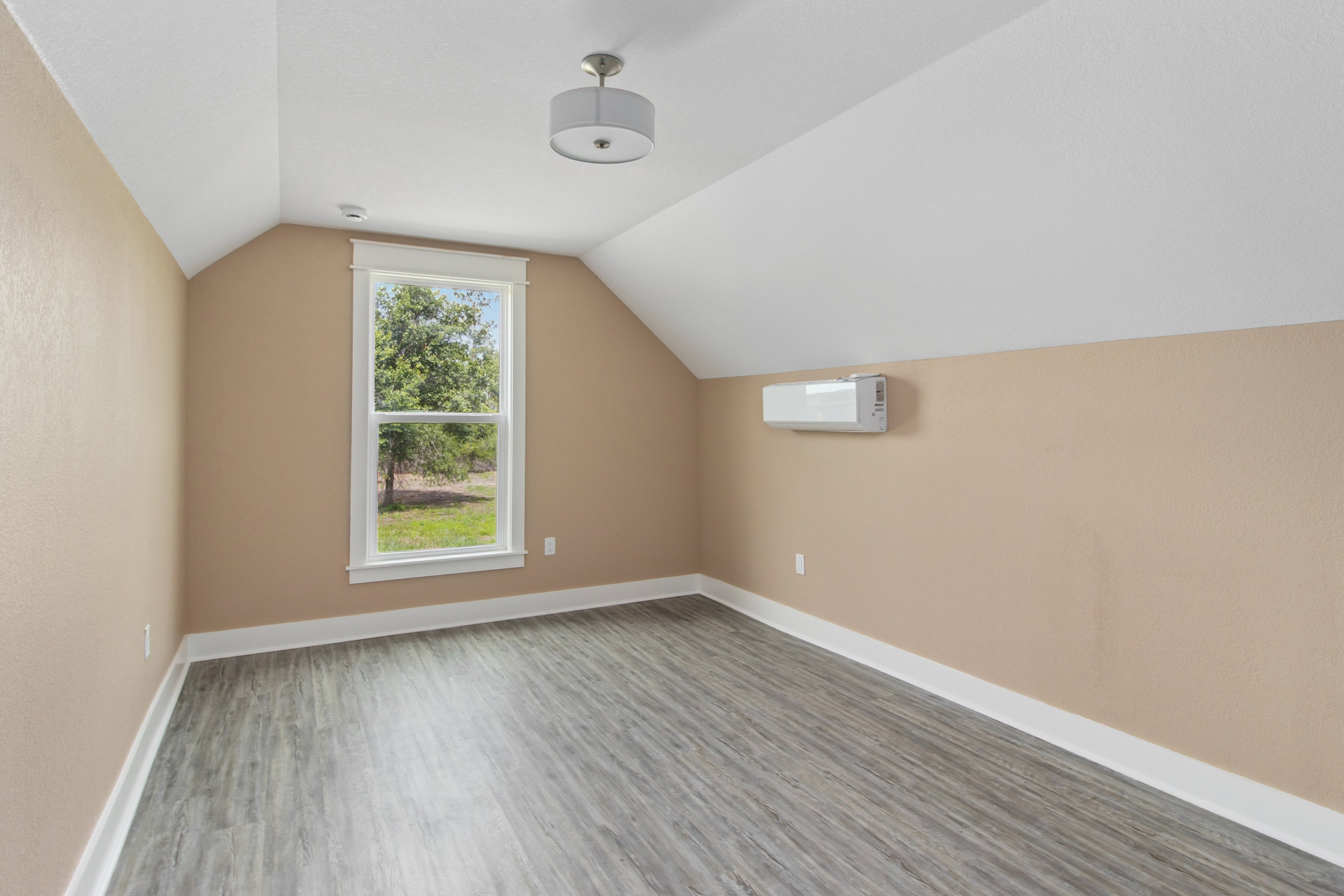 Sunlit room with wide window overlooking trees, light wood flooring, white walls, modern ceiling light fixture, and simple baseboard molding.