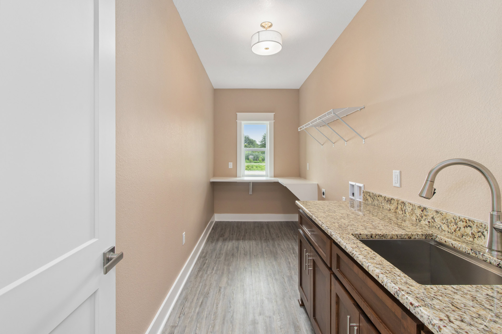 White kitchen counter with stainless steel sink beneath a window showing trees outside, white tile backsplash, floating white shelf, and modern light fixture with white shade.