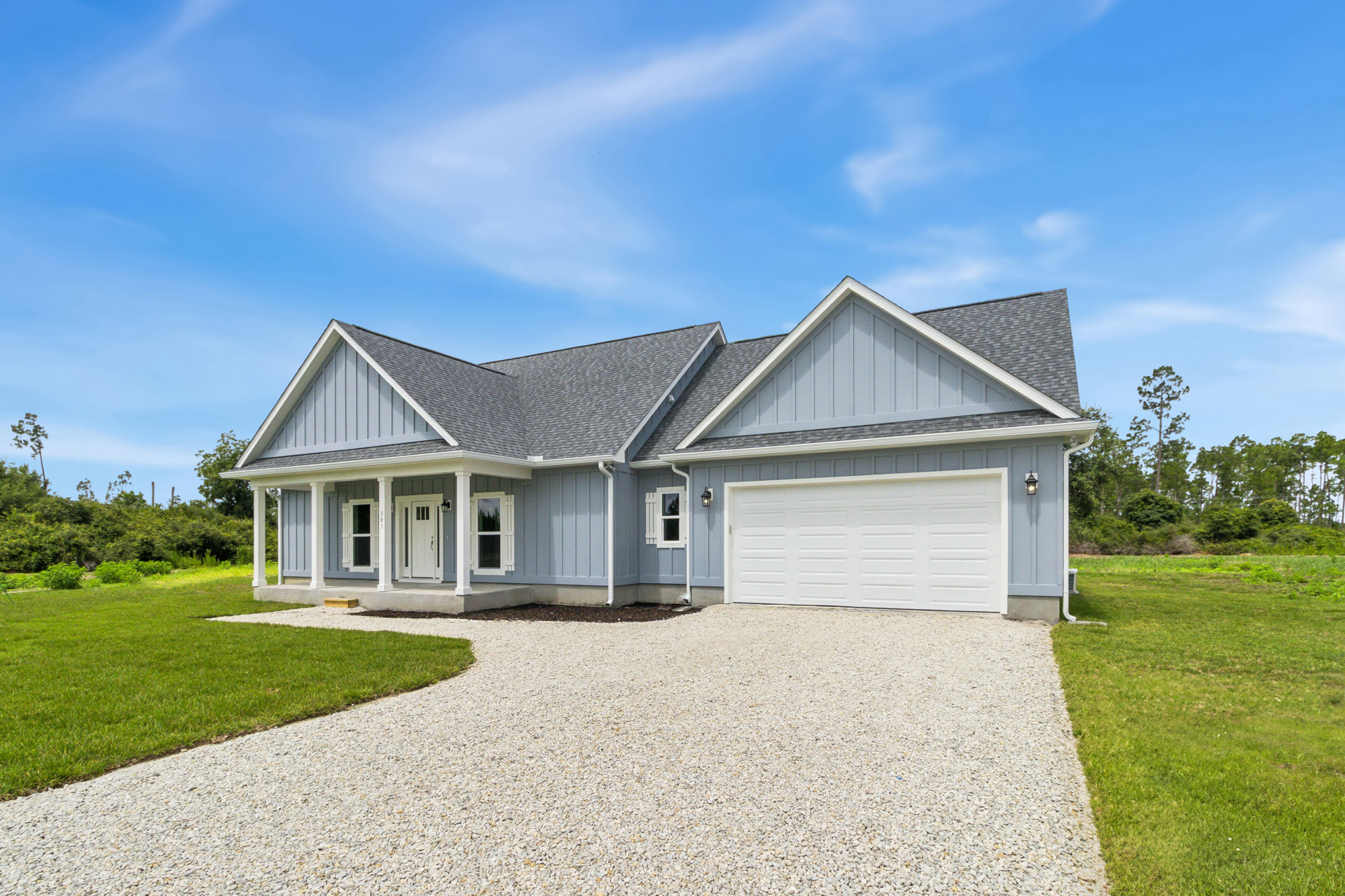Two-story house with gray siding, white garage door, gravel driveway bordered by grass, blue sky overhead, Robert Frost Farm visible in the background.