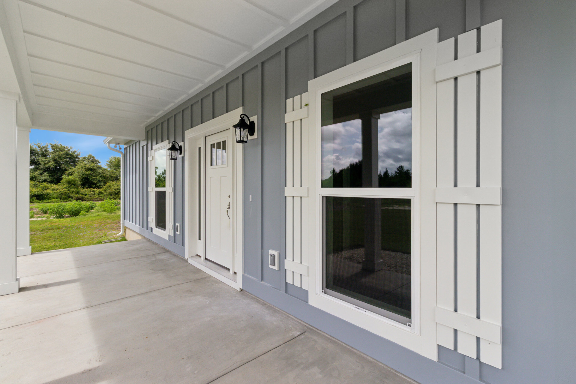 Two-story home with wide front porch, white railings, multiple windows, concrete flooring, and trees reflected in glass under blue sky