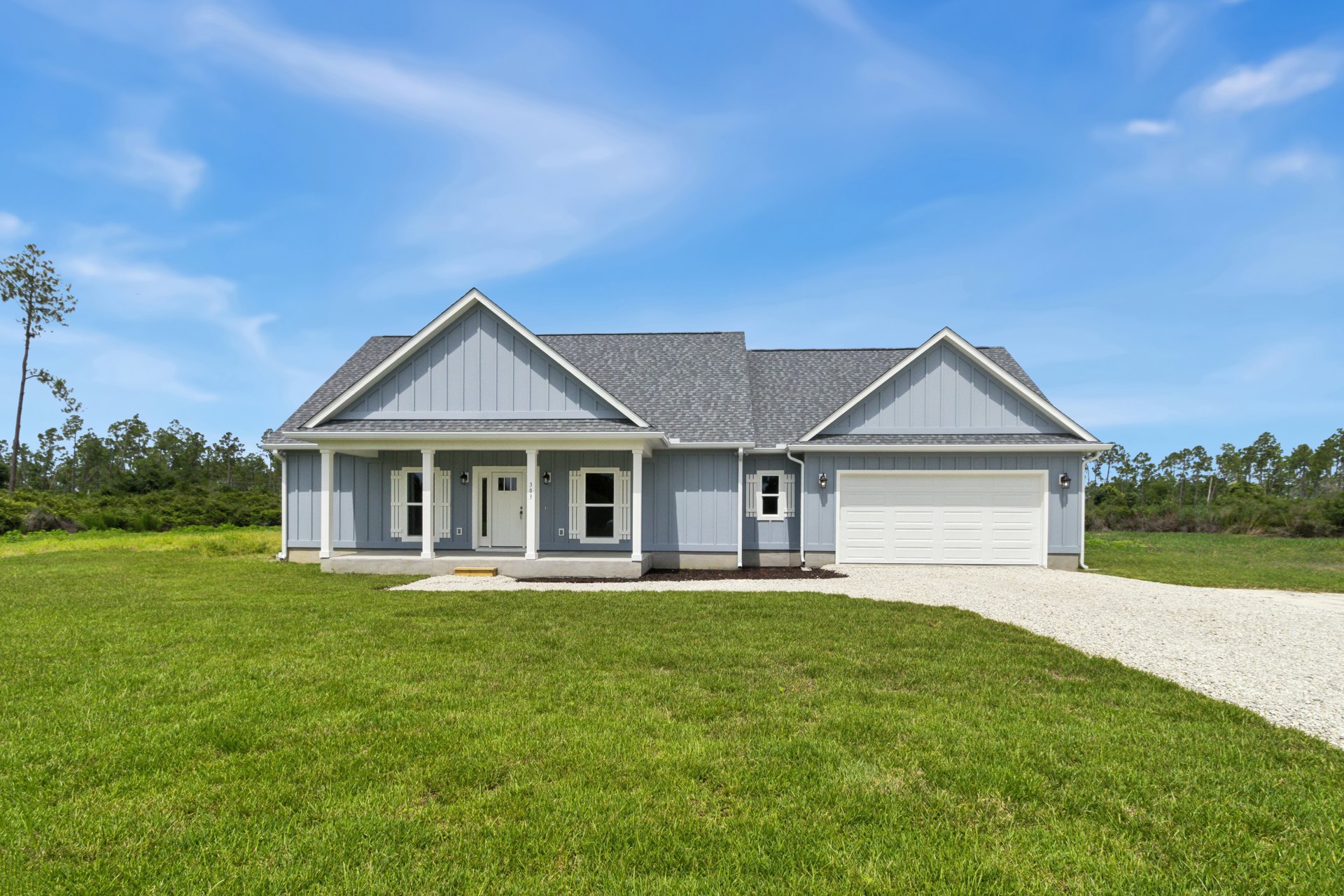 Two-story house with grey roof, white-framed windows, and covered porch, set behind a green lawn with gravel edging; tree and blue sky with clouds in background