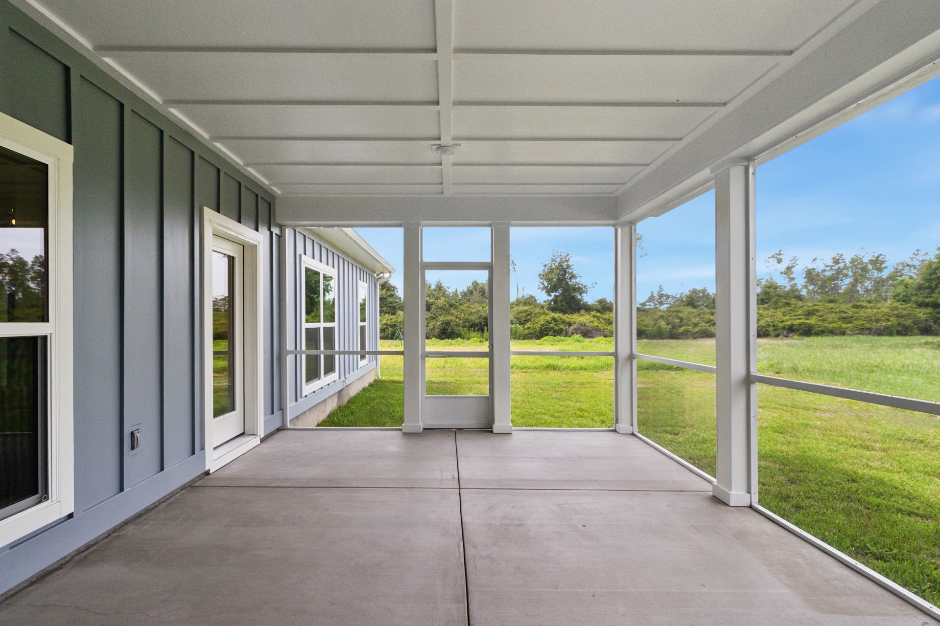 Covered porch with white ceiling and light fixture, concrete patio floor, white-framed windows and door, overlooking green grass and trees