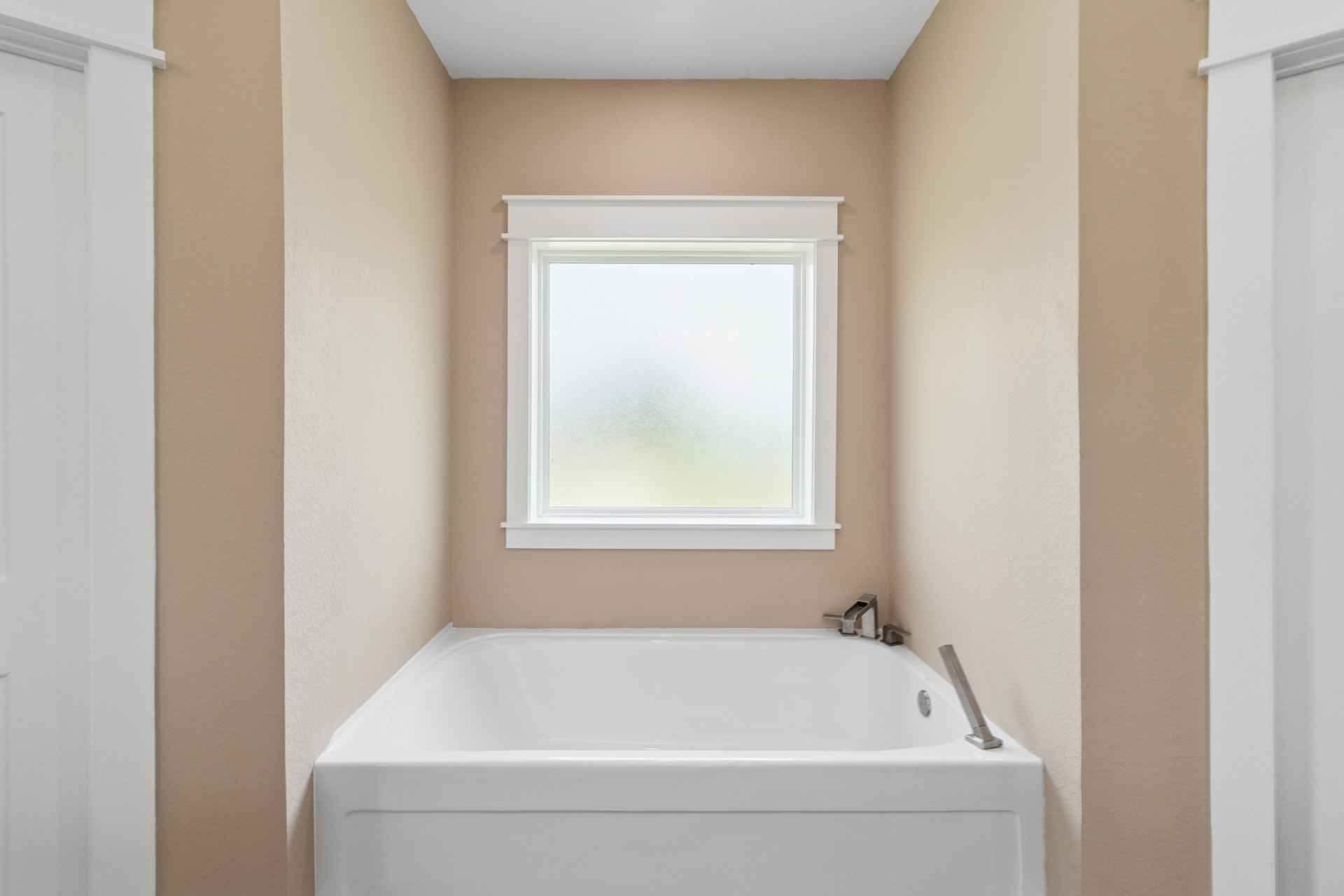 Freestanding white bathtub on white tile floor, frosted glass window above, chrome towel bar mounted on white wall with brown wood accent trim