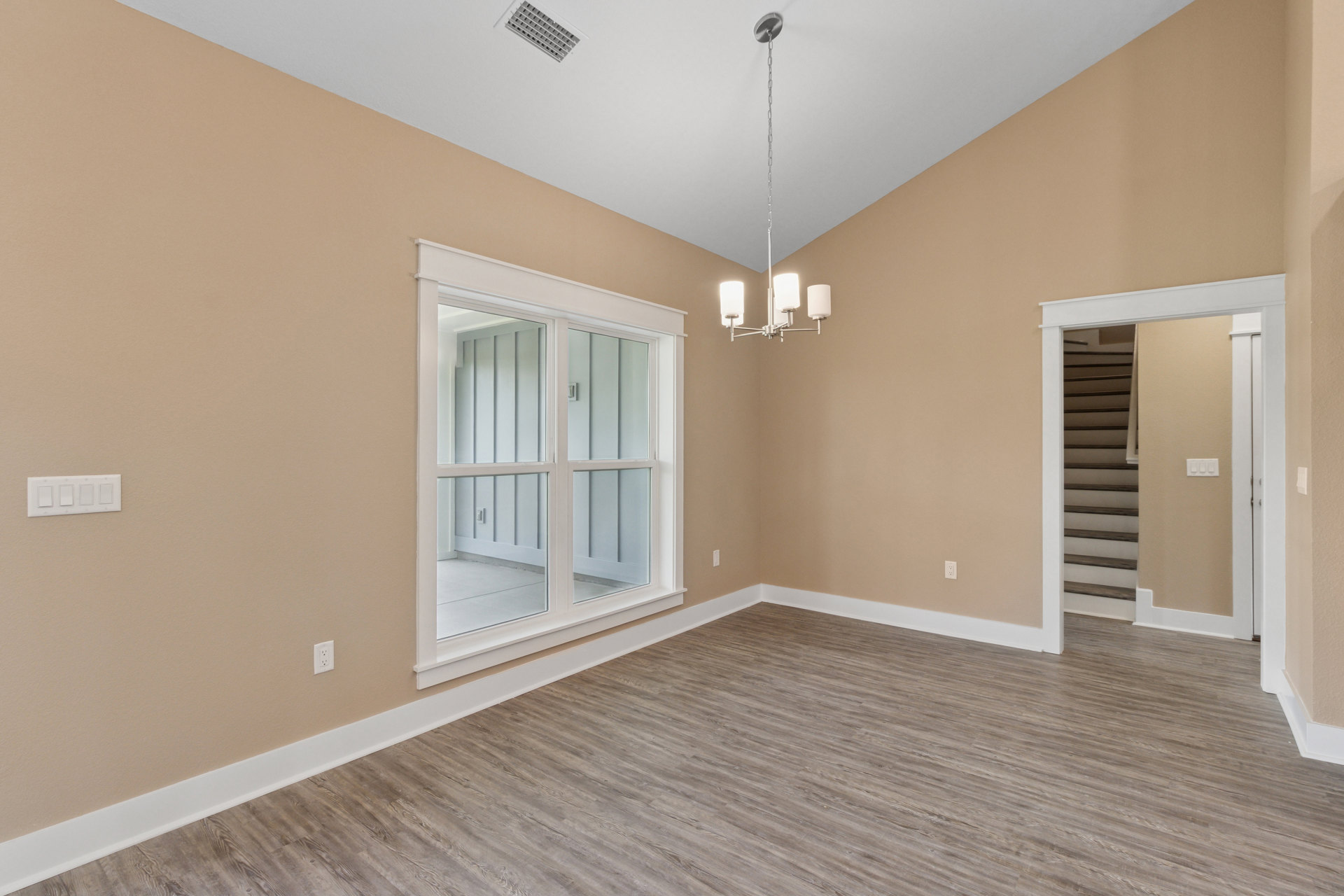 Bright room featuring wood laminate flooring, white plaster walls, large window with glass panes, ceiling vent, and doorway leading to staircase with painted railing.