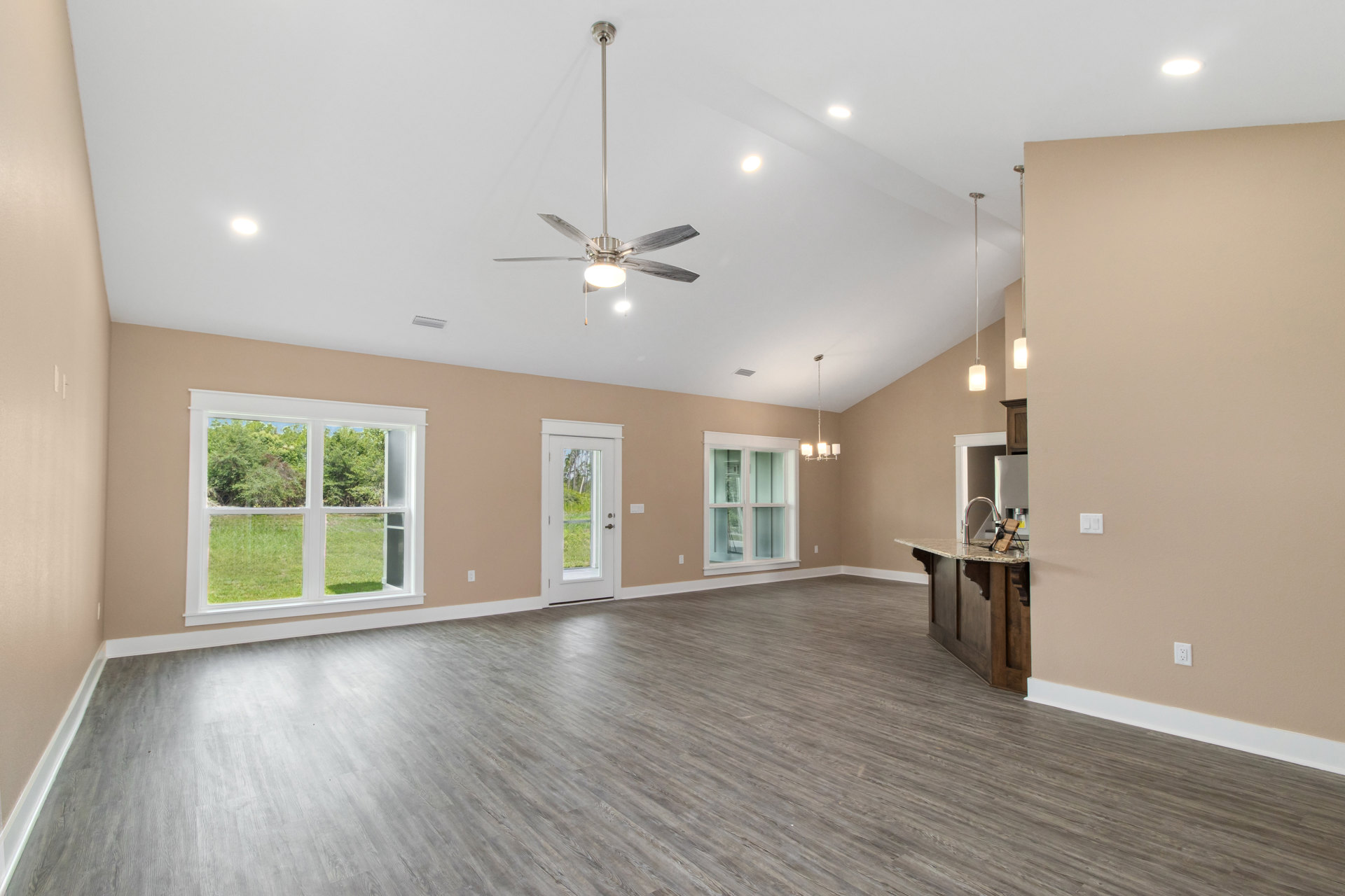 Spacious living room featuring wood flooring, ceiling fan with light fixture, multiple windows with views of trees and forest, and a door with glass panes.