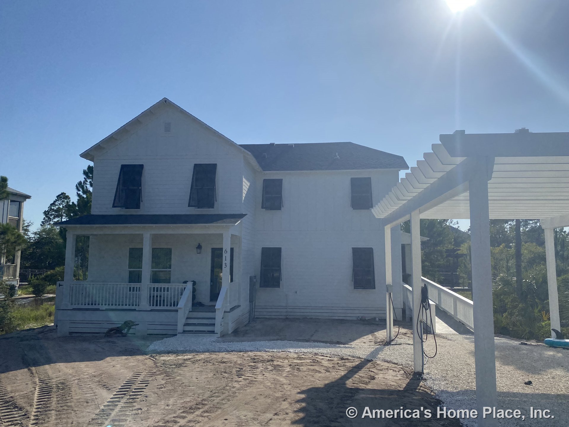 Two-story white house with black shutters, lap siding exterior, covered front porch with white railing, white pergola structure, gravel driveway, and prominent entry door.