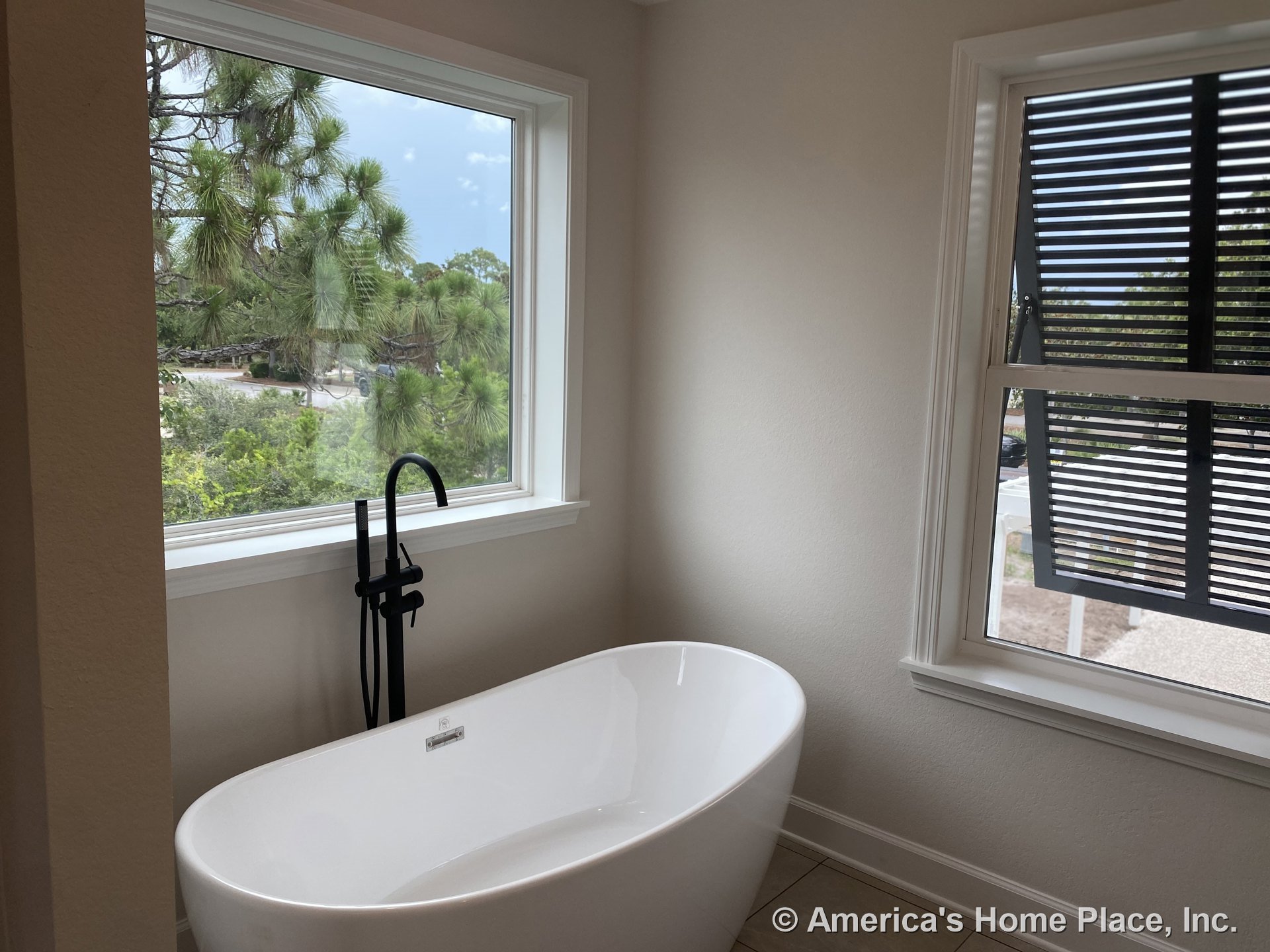 Freestanding bathtub with matte black floor-mounted faucet positioned near two large picture windows with white trim, tile flooring, partial window shutters, and neutral wall