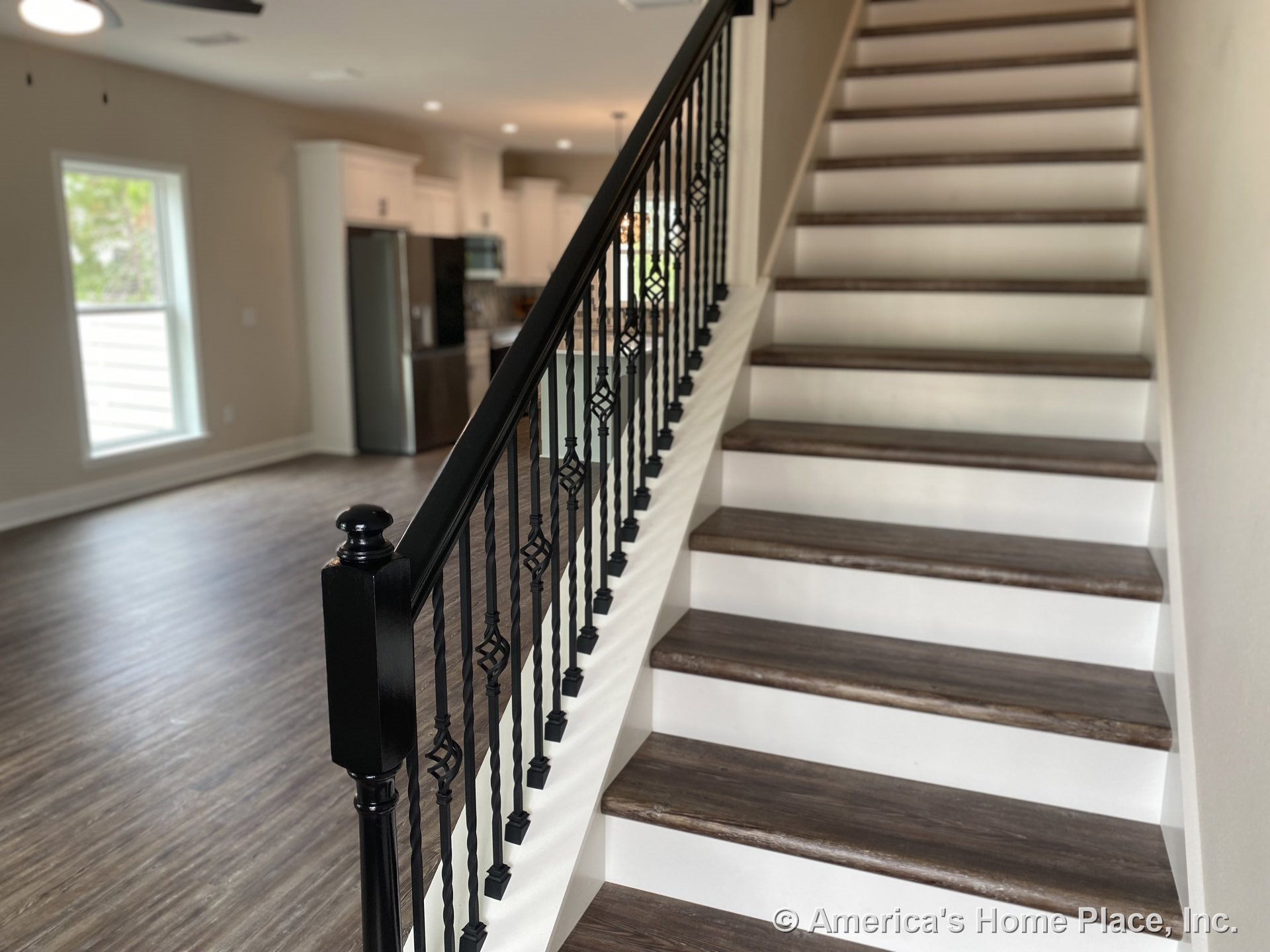 Staircase with dark wood treads and black iron balusters in open-plan interior featuring wood-look plank flooring, large window with white trim, white kitchen cabinetry, stainless
