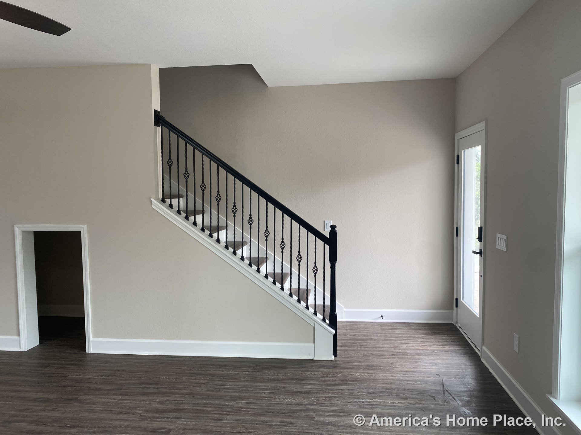 Black iron stair railing with wood-look plank flooring, glass exterior door, white trim and baseboards, small under-stair doorway, and neutral wall paint in a modern living area.