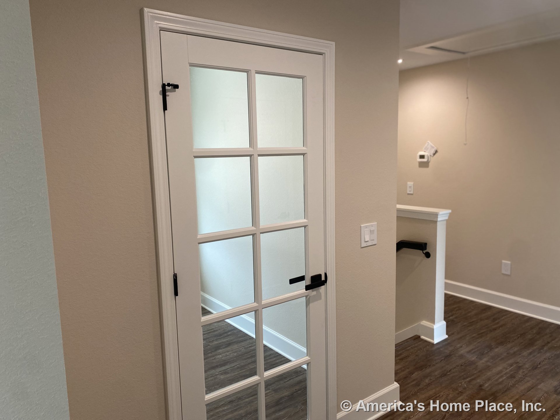 Glass-paneled interior door with white trim dividing two rooms featuring wood-look flooring, beige painted walls, baseboard molding, wall-mounted thermostat, light switch, and