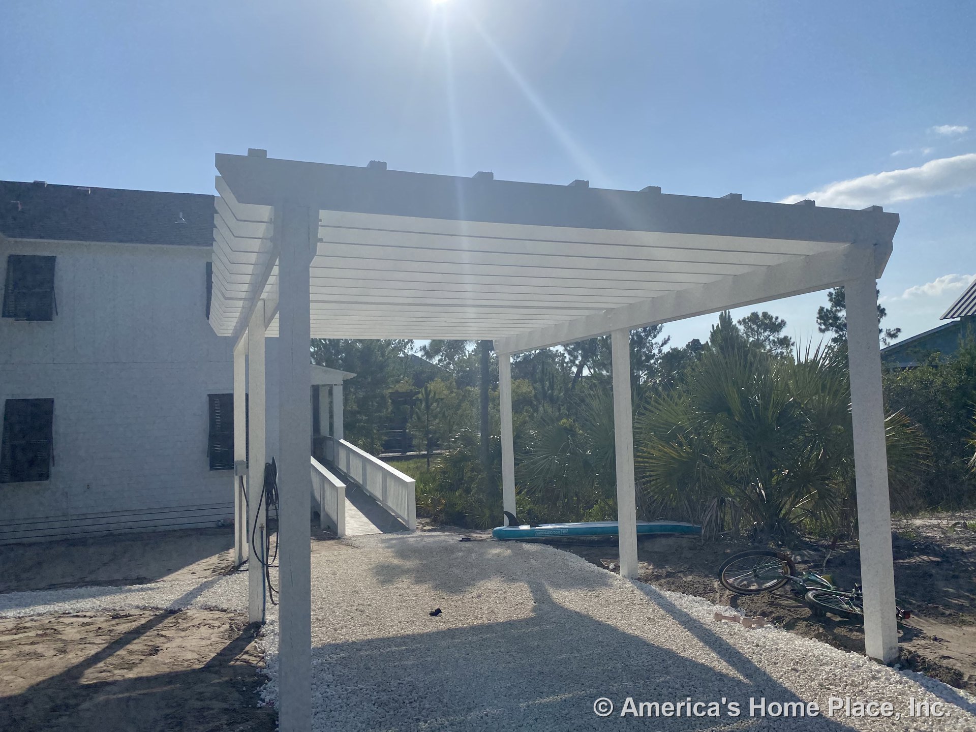 White pergola with gravel flooring attached to house exterior, featuring ramp with metal railing, black window shutters, and white trim under bright sunlight.