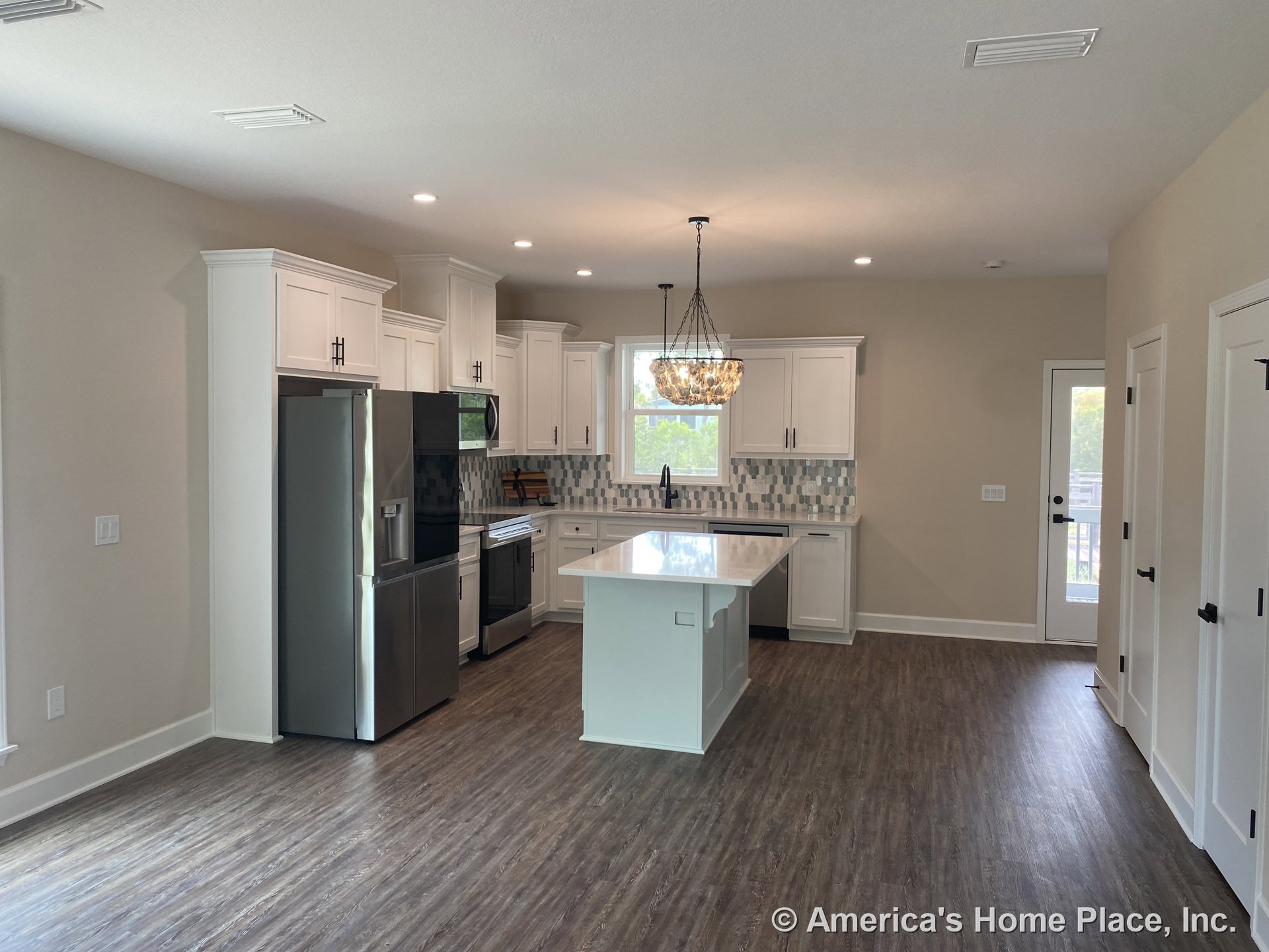 White shaker cabinets and stainless steel appliances surround a central island with quartz countertop, geometric tile backsplash, pendant chandelier lighting, wood-look vinyl
