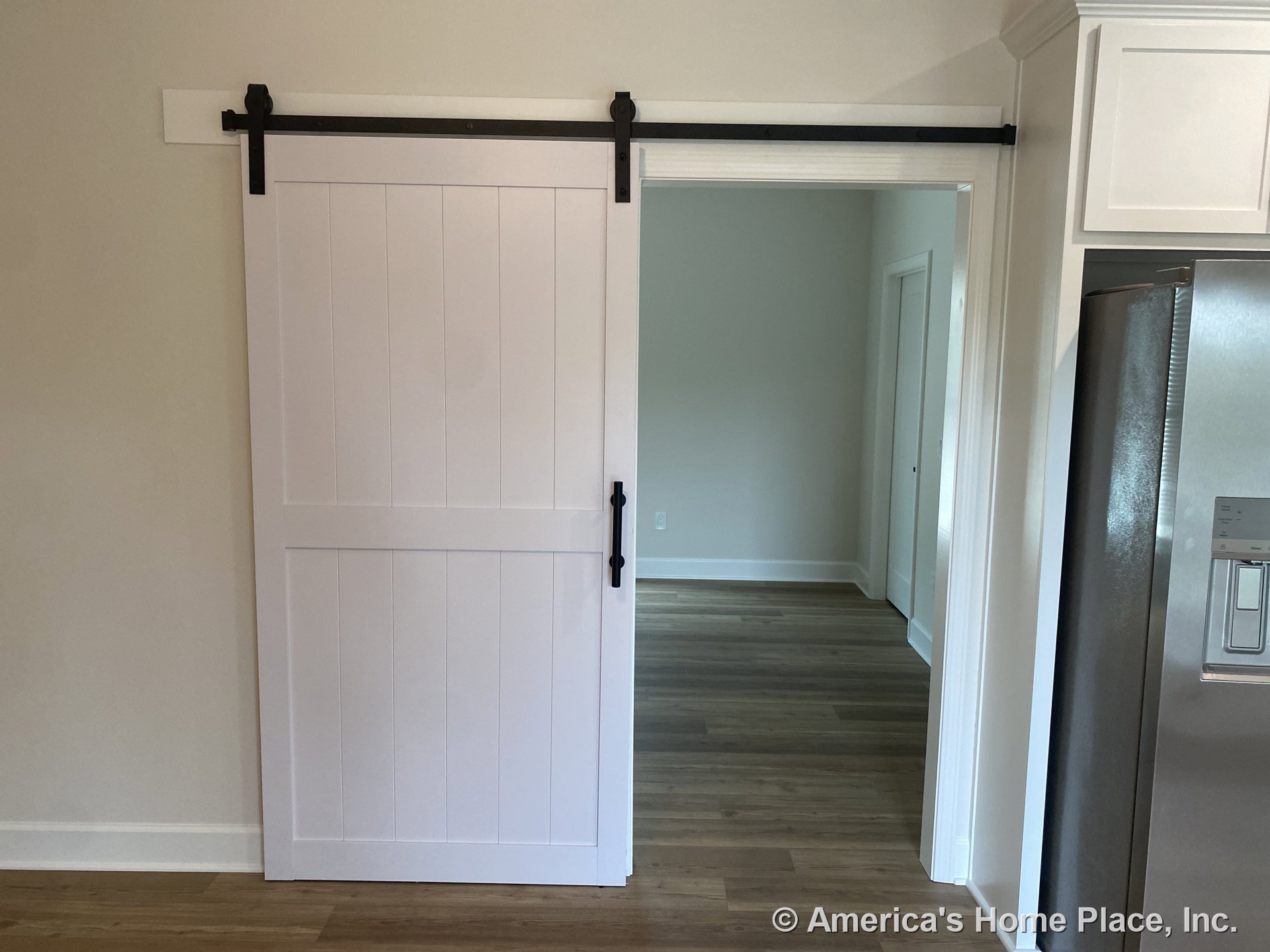 White sliding barn door with black hardware dividing kitchen and adjacent room; wood plank flooring throughout, white cabinetry, stainless steel refrigerator, and trimmed doorway