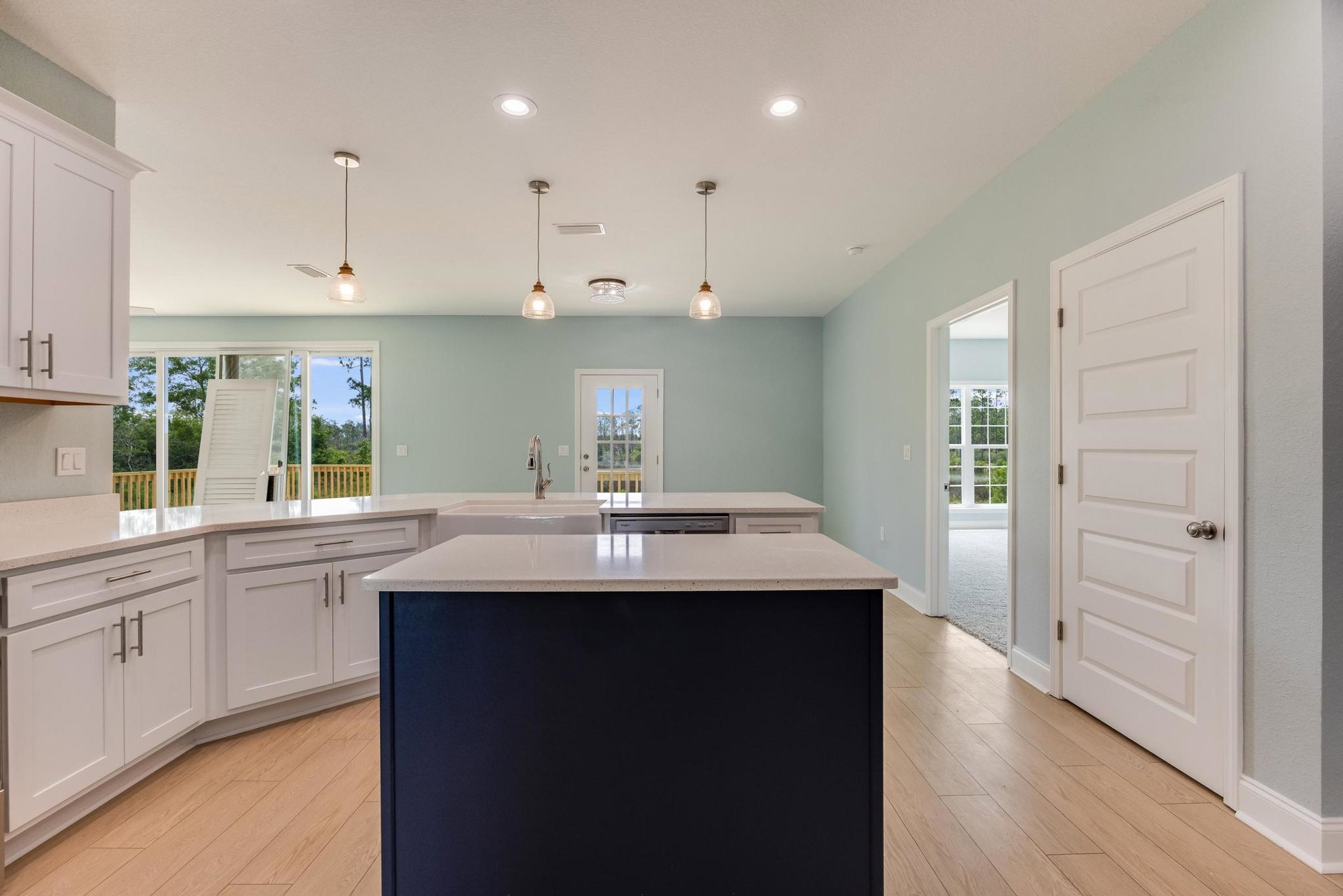 Open kitchen with white island countertop, pendant light fixtures, tile backsplash, white cabinetry, stainless steel sink, and glass-paneled door with silver knob