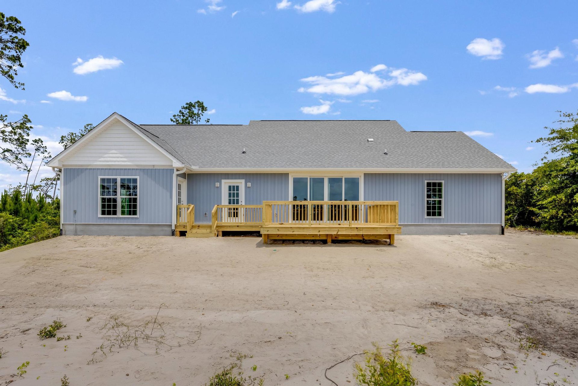 Two-story home with white siding, multi-pane windows, glass-paneled door, wooden deck with railing, covered porch, sandy yard, and surrounding trees under a partly cloudy sky