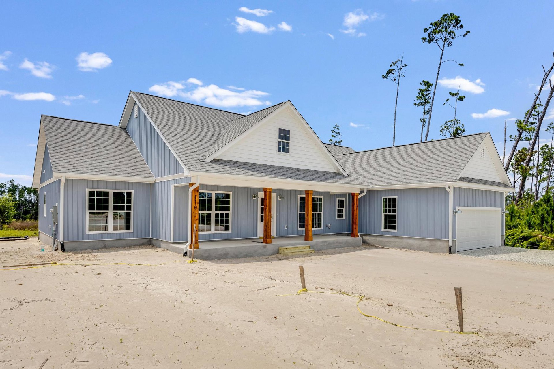 Two-story home with blue roof, white-framed windows, and sandy driveway; yellow caution tape on wooden post near unfinished landscaping.