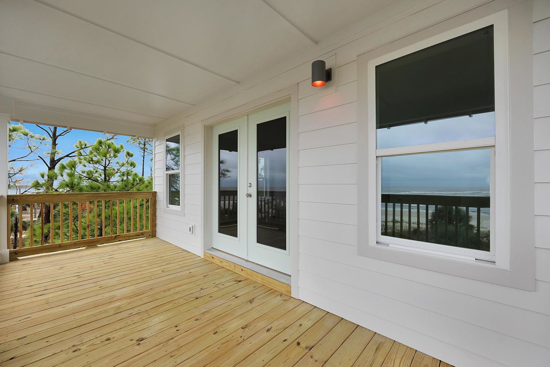 Wood deck with wooden railing, glass door, and large windows overlooking trees and ocean; black outdoor lamp with warm orange light.