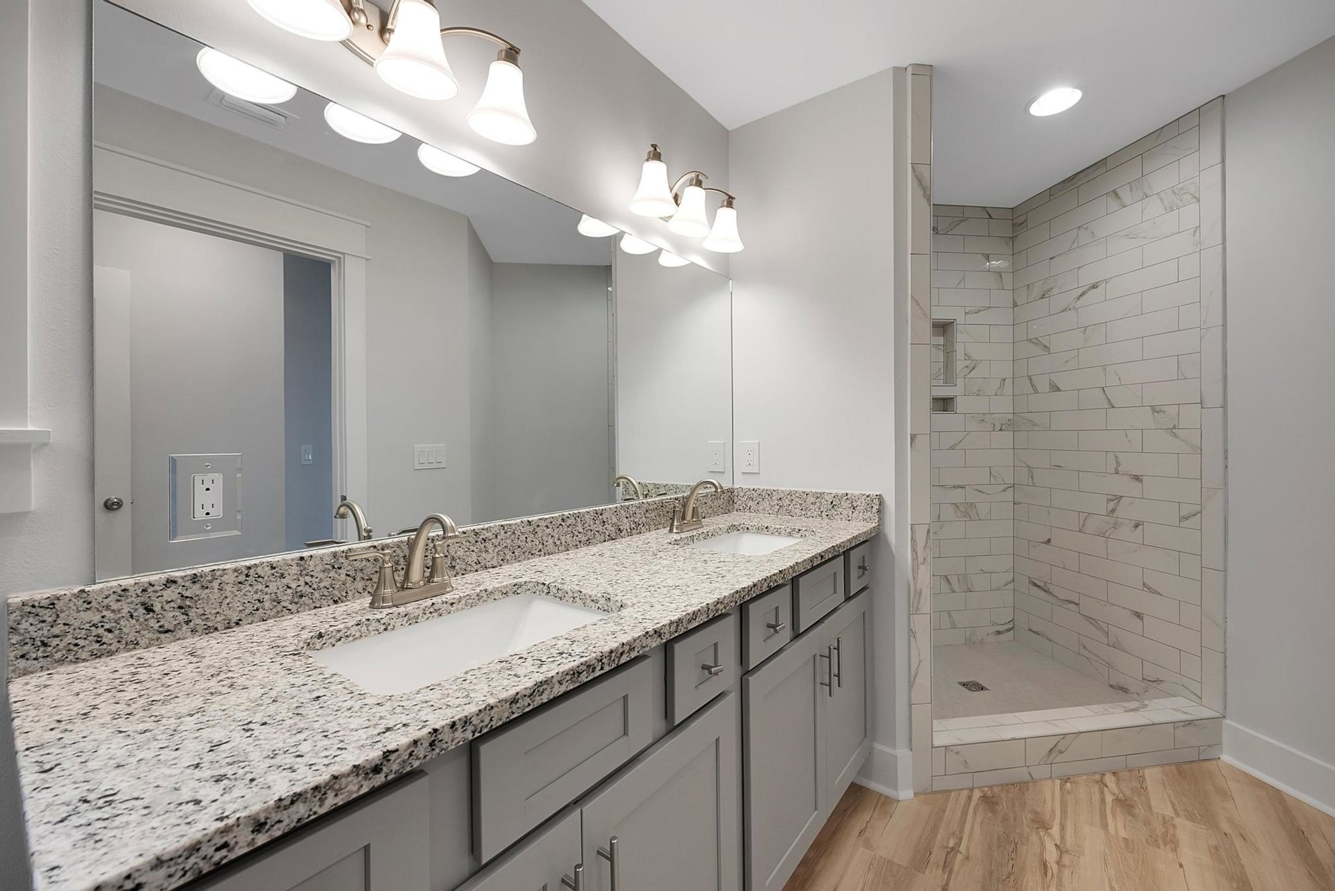 Bathroom with expansive mirror above marble countertops, undermount sink, chrome faucet, tile shower wall, recessed ceiling light, and white cabinetry.